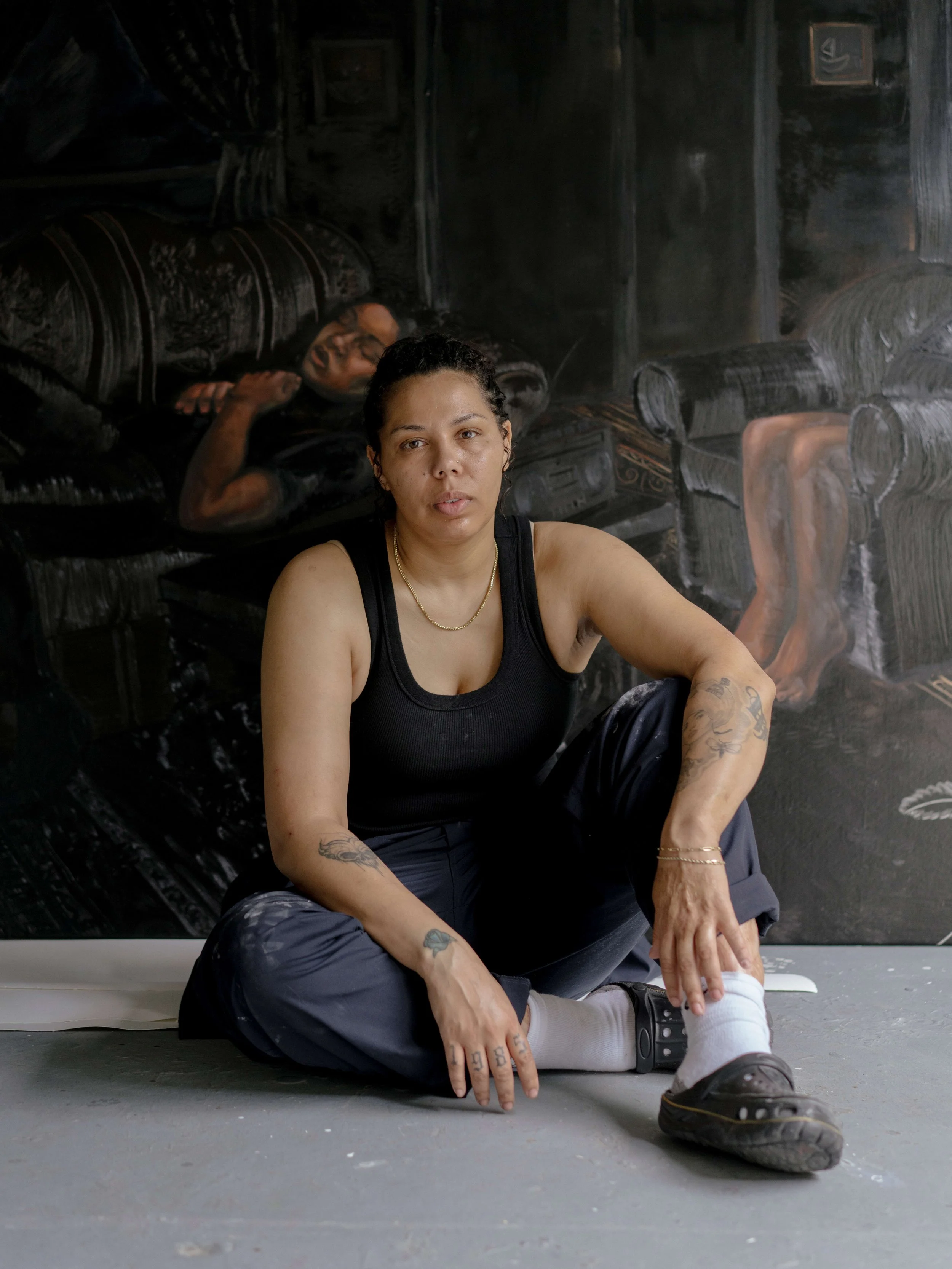 Portrait of artist Natia Lemay seated in her Toronto studio in front of a large figurative painting in all black.