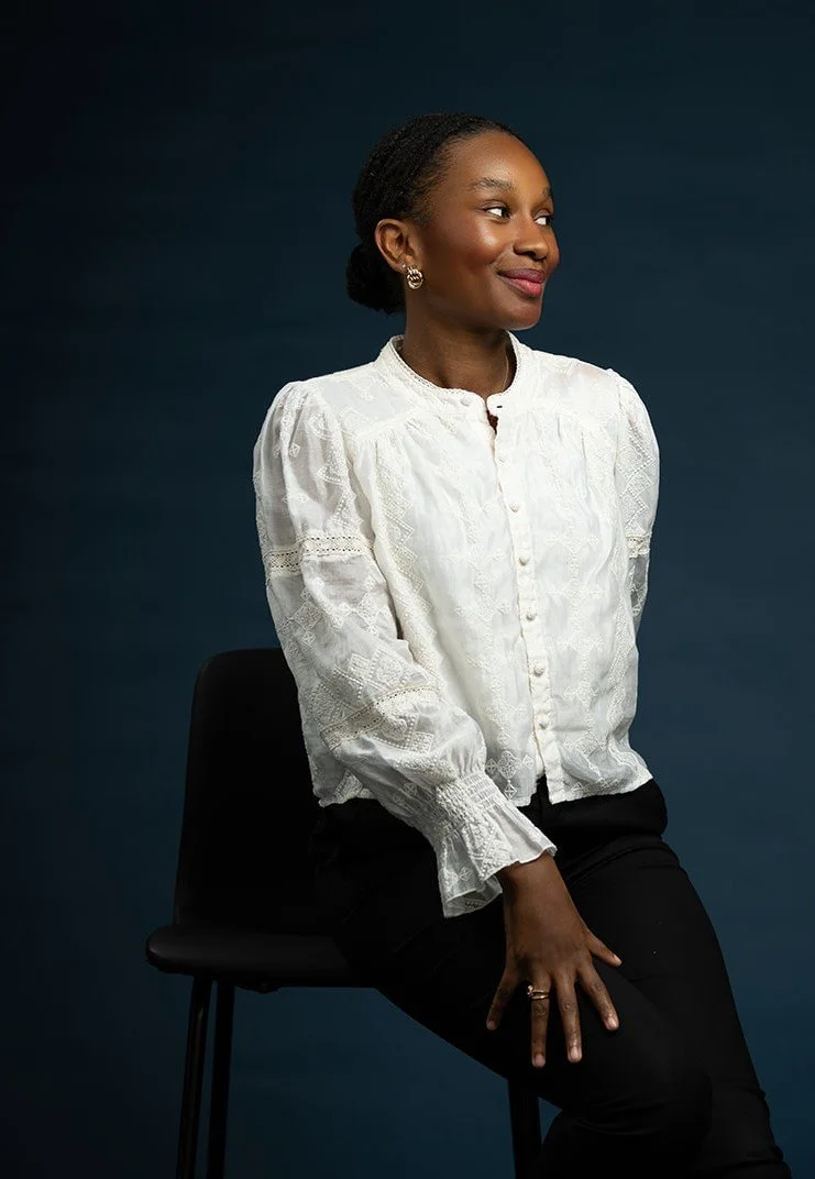 A woman with dark skin, short hair, wearing a white embroidered blouse and black pants, sitting on a black chair against a dark blue background, looking to her right with a slight smile.