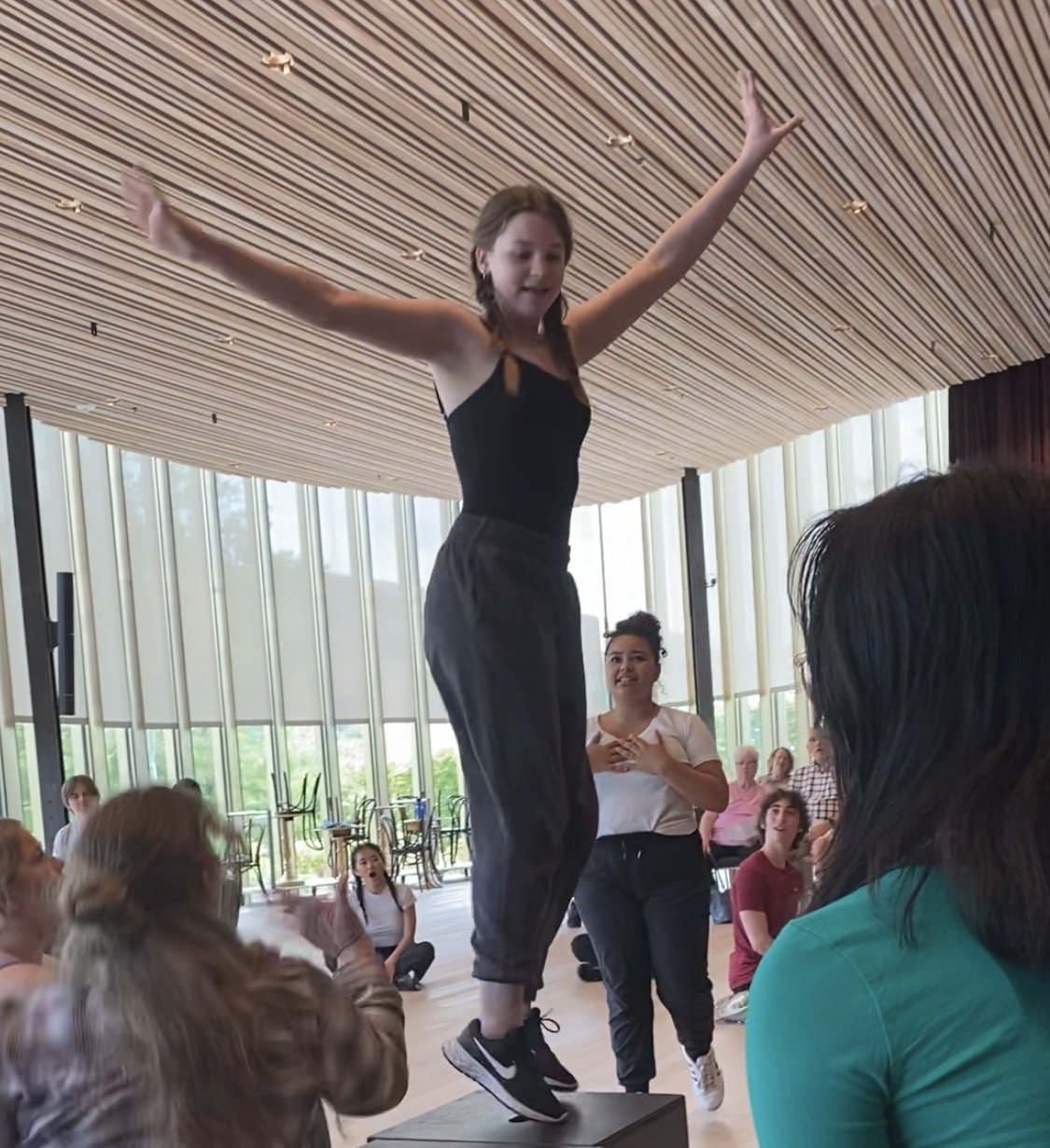 A young woman stands on a raised platform with arms outstretched, surrounded by a group of people indoors. Large windows with curtains are in the background, and some people are sitting or standing, watching her.