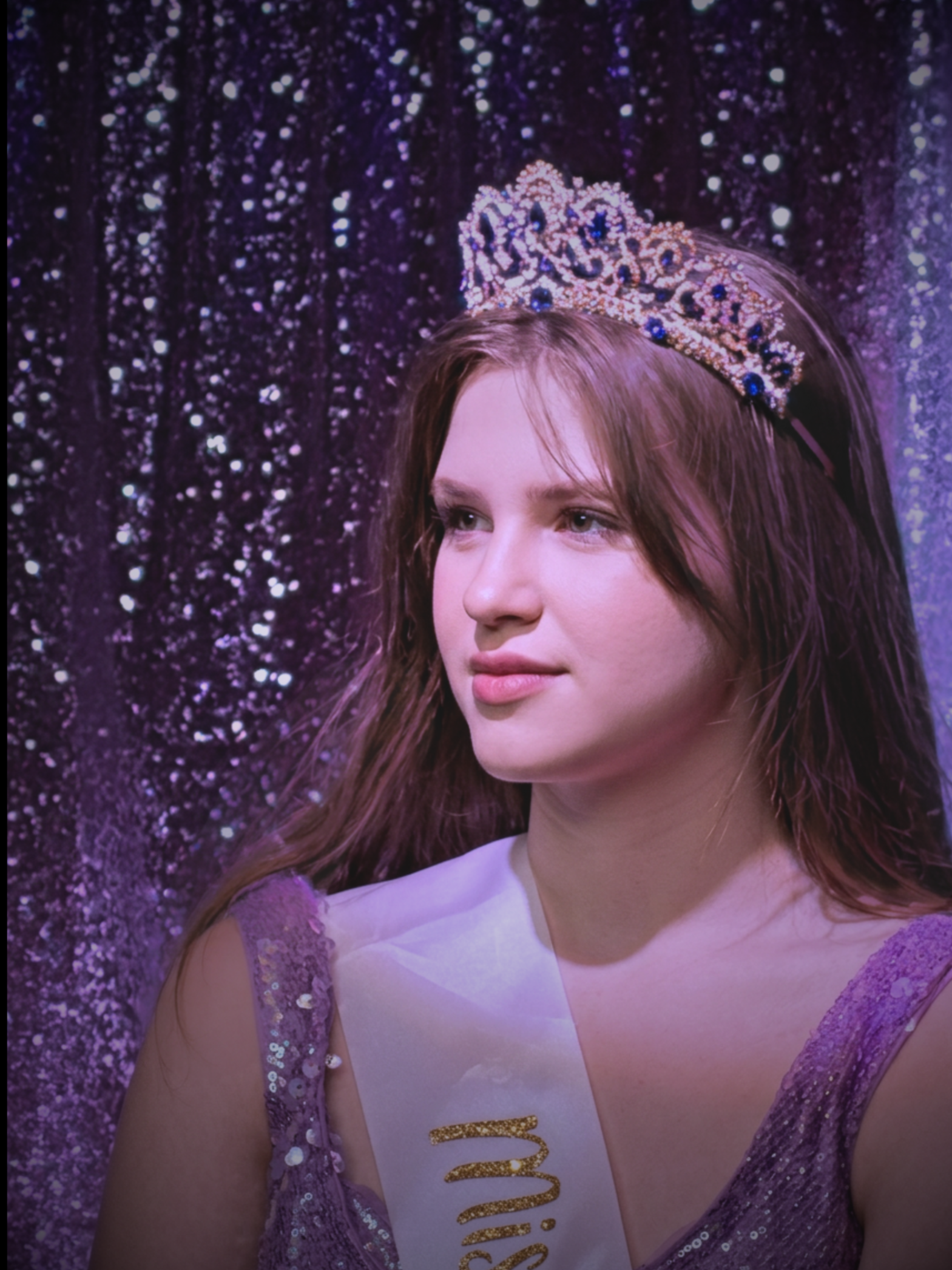 Young woman wearing a tiara and sash, standing in front of a glittery purple curtain, celebrating a pageant or beauty contest.