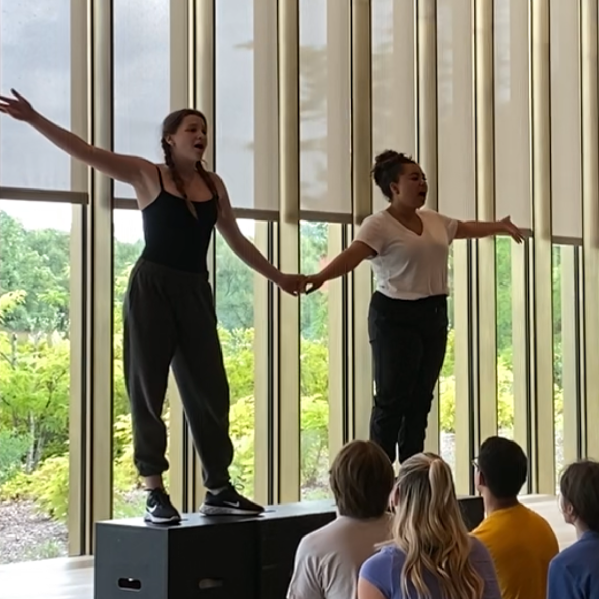 Two women perform a dance or theatrical act on a raised platform, holding hands with arms outstretched, in front of a large window with a view of green trees, as an audience watches.
