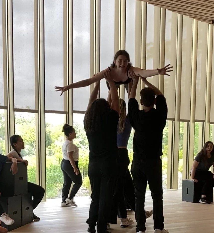 A girl being lifted by several people in a cheerleading or dance practice, with others observing inside a room with large glass windows showing greenery outside.