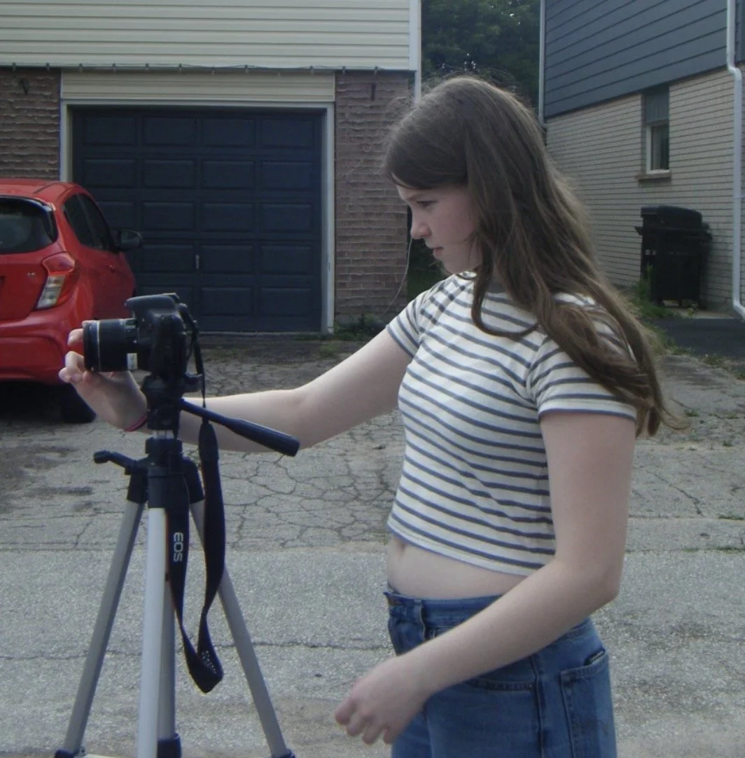 A young woman with long brown hair wearing a striped crop top and jeans, adjusting a camera on a tripod outdoors in a residential driveway.