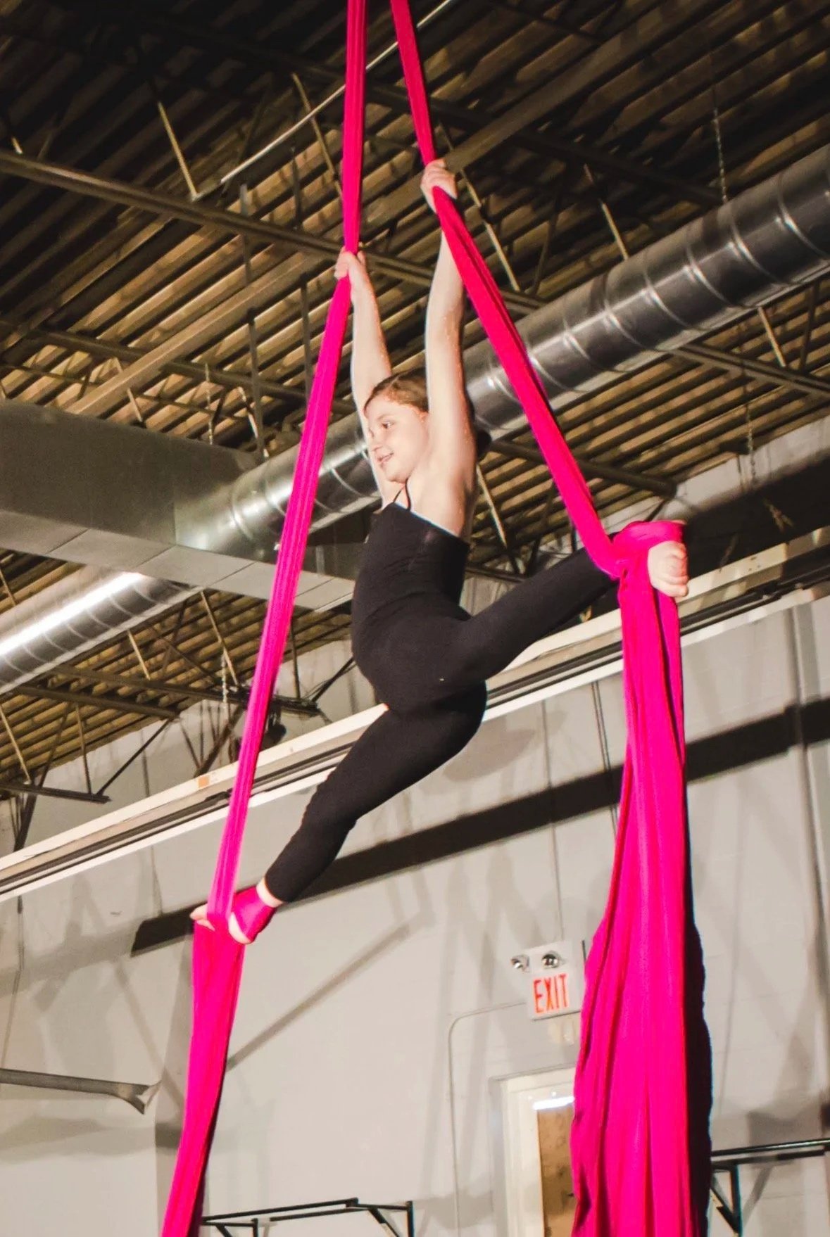 A young woman practicing aerial silk acrobatics in a gym, hanging from pink fabrics with her arms raised and one leg extended.