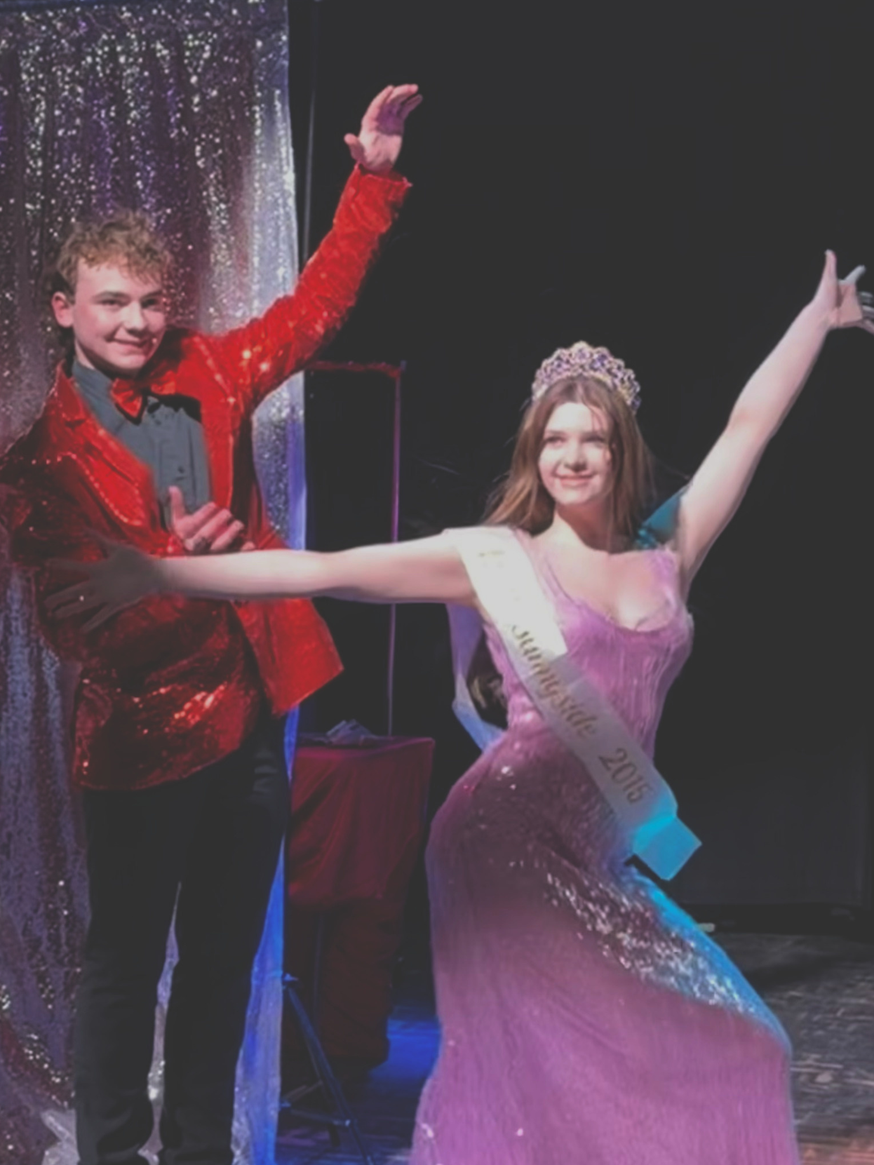 A young man in a red suit and a young woman in a pink dress and crown, with a sash, posing on stage after a pageant.