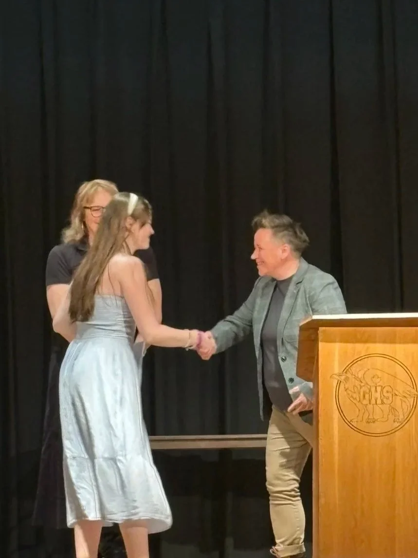 Woman in gray dress shaking hands with woman in gray blazer and black shirt on stage with two other women, black curtain backdrop, wooden podium to the right, school logo visible.