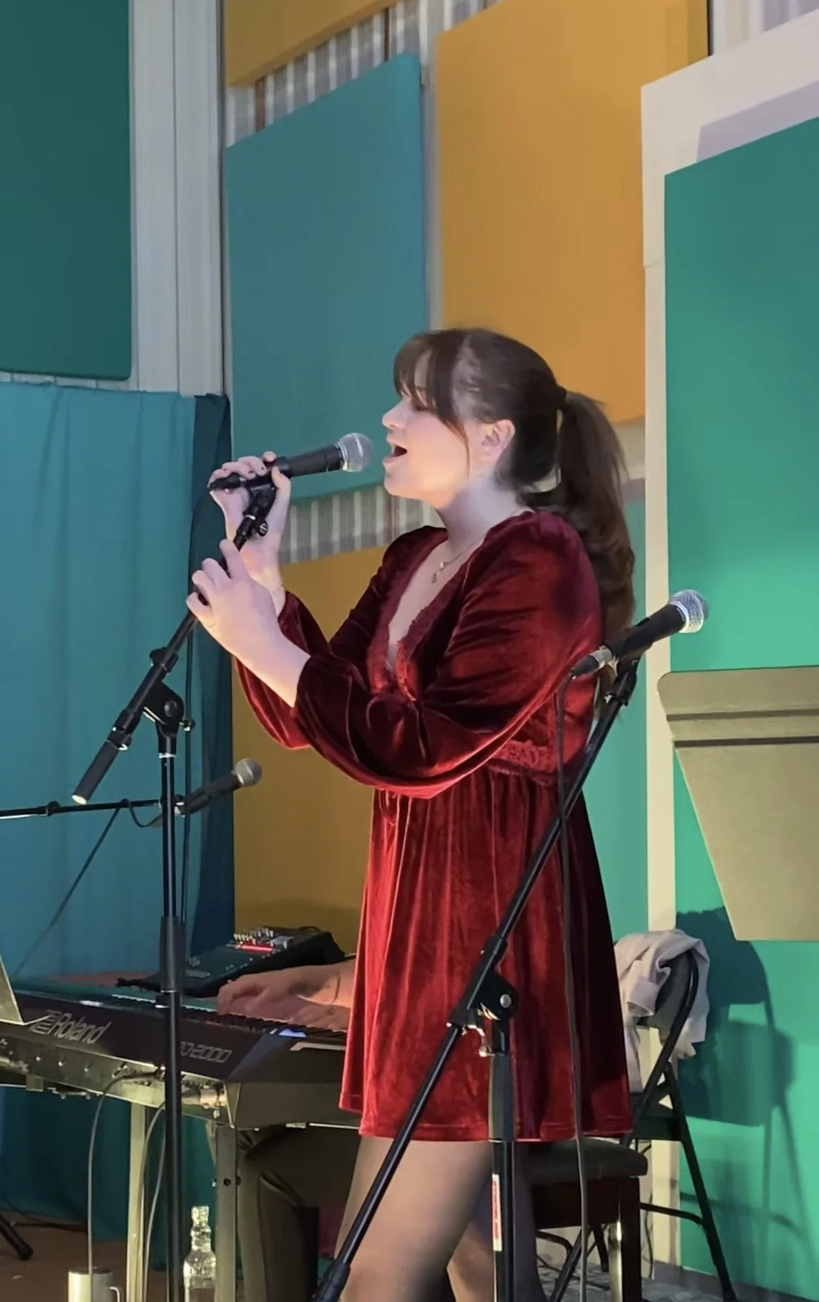 A woman wearing a red velvet dress singing into a microphone, playing a keyboard on stage with colorful panels in the background.