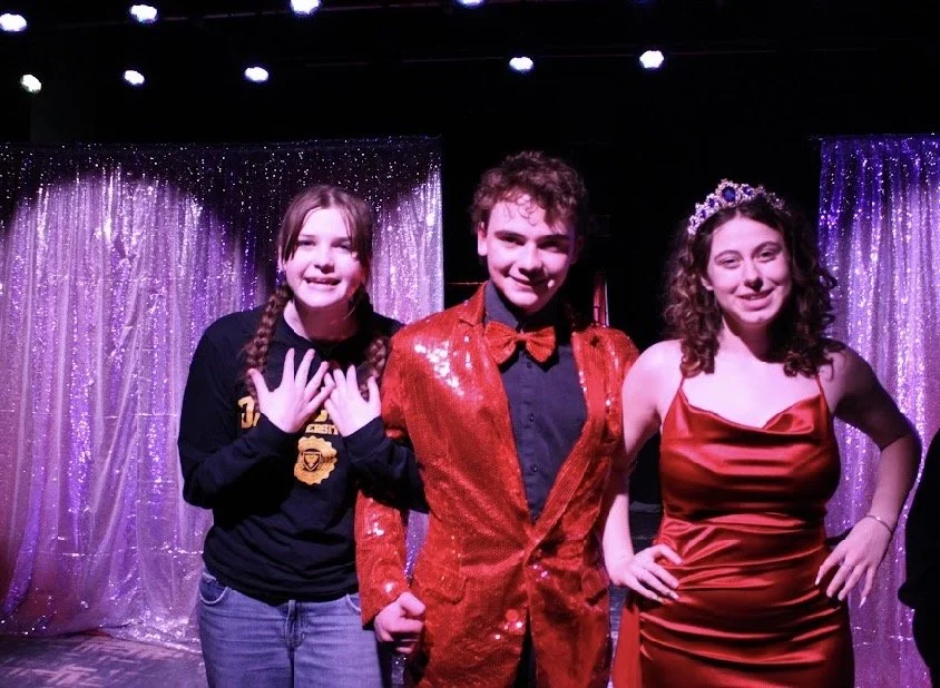 Three people posing on stage with shiny purple curtains in the background. One girl on the left is wearing a black hoodie with yellow graphics, the boy in the middle is dressed in a red sequined blazer and red bow tie, and the girl on the right is wearing a red satin dress and a tiara.