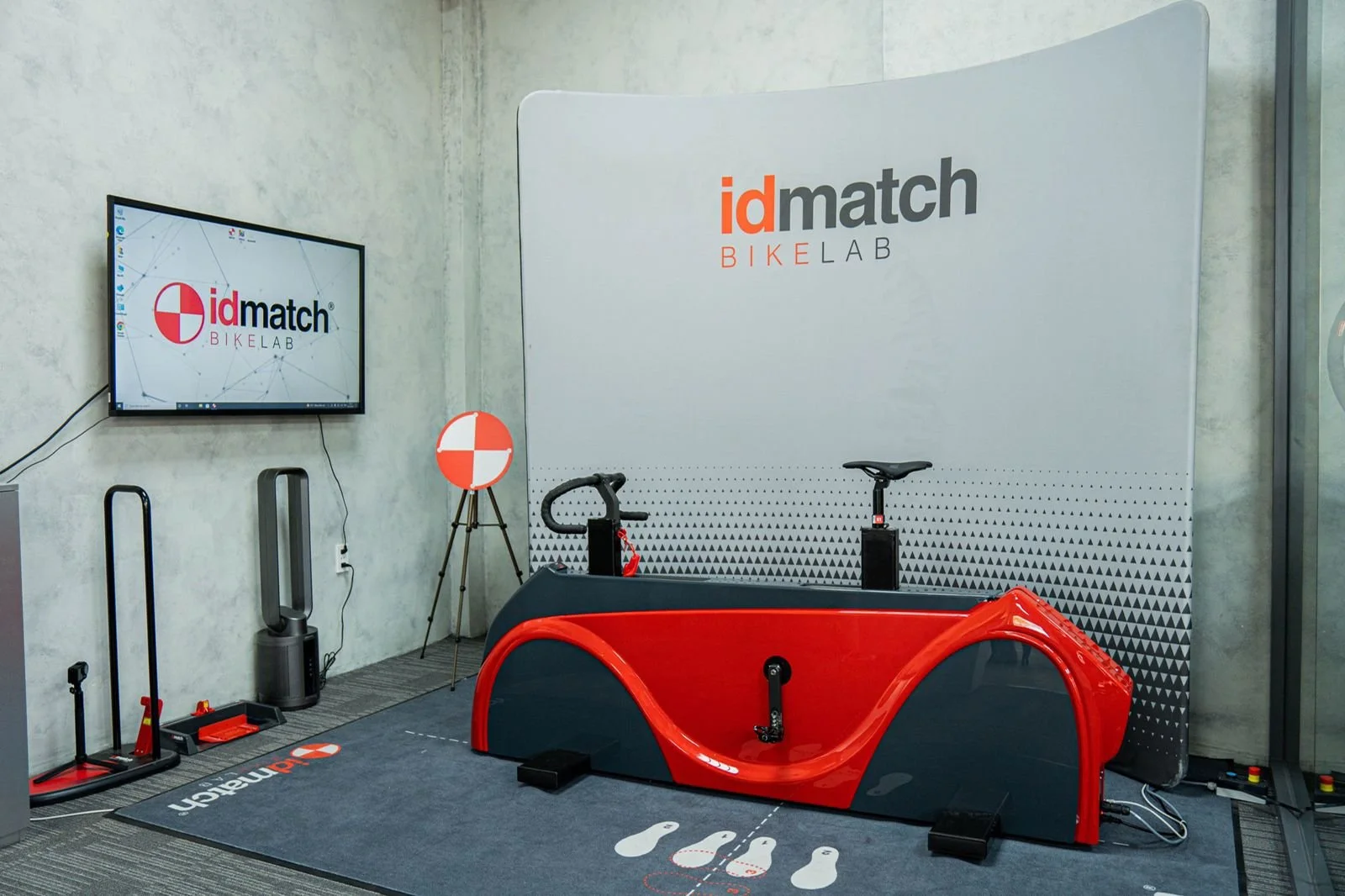 Indoor gym or bike lab with a red and black stationary cycling trainer, a wall display with 'idmatch BIKE LAB' logo, a TV monitor showing the same logo, fitness equipment, and a gray wall.