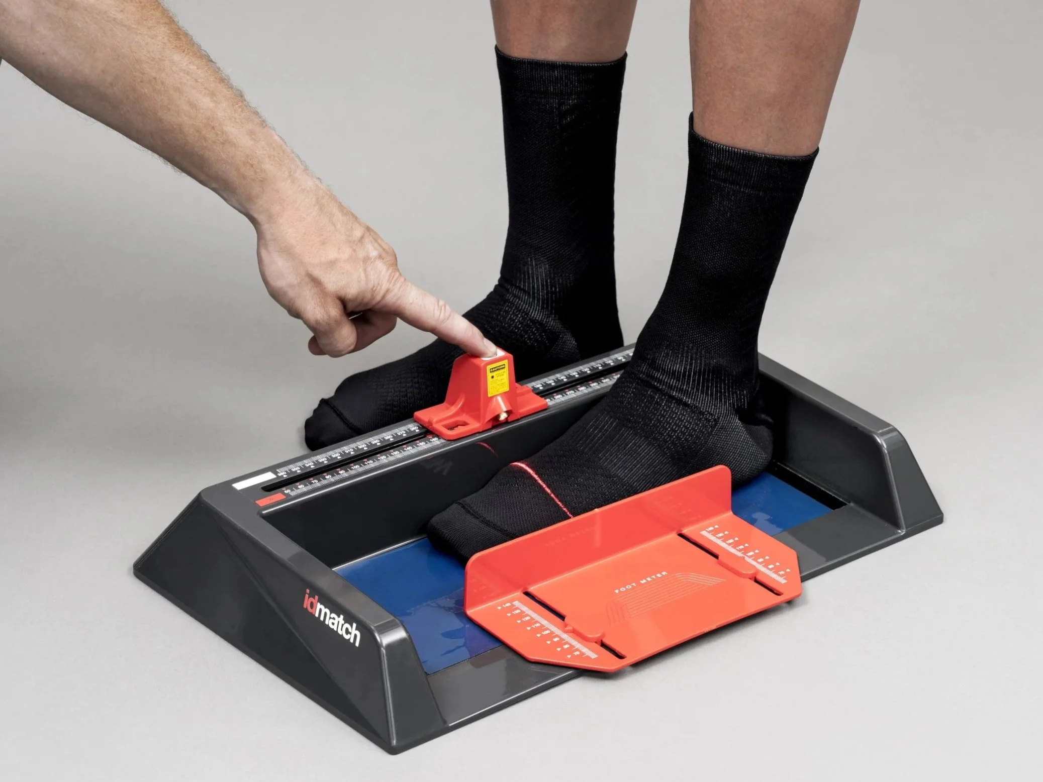 Two people using a digital foot measuring device, with one pointing at a red calibration tool on the device and the other standing on it wearing black socks.