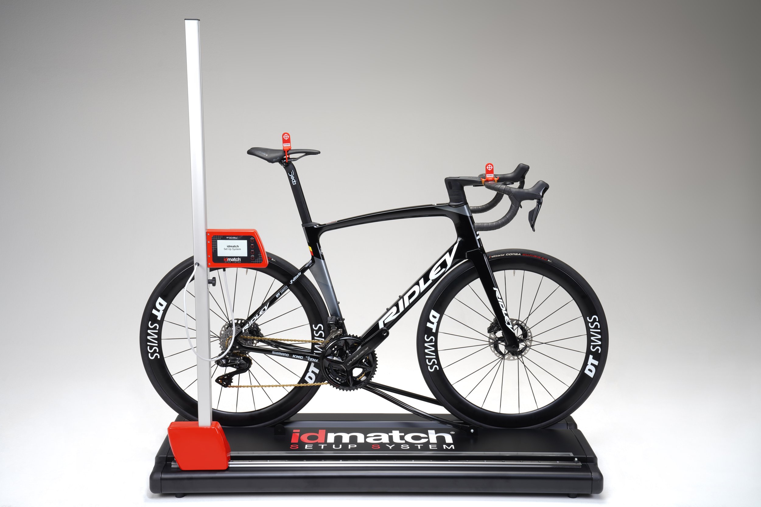 A black road bicycle mounted on an indoor bike fitting system on a black platform, with a gray wall background.