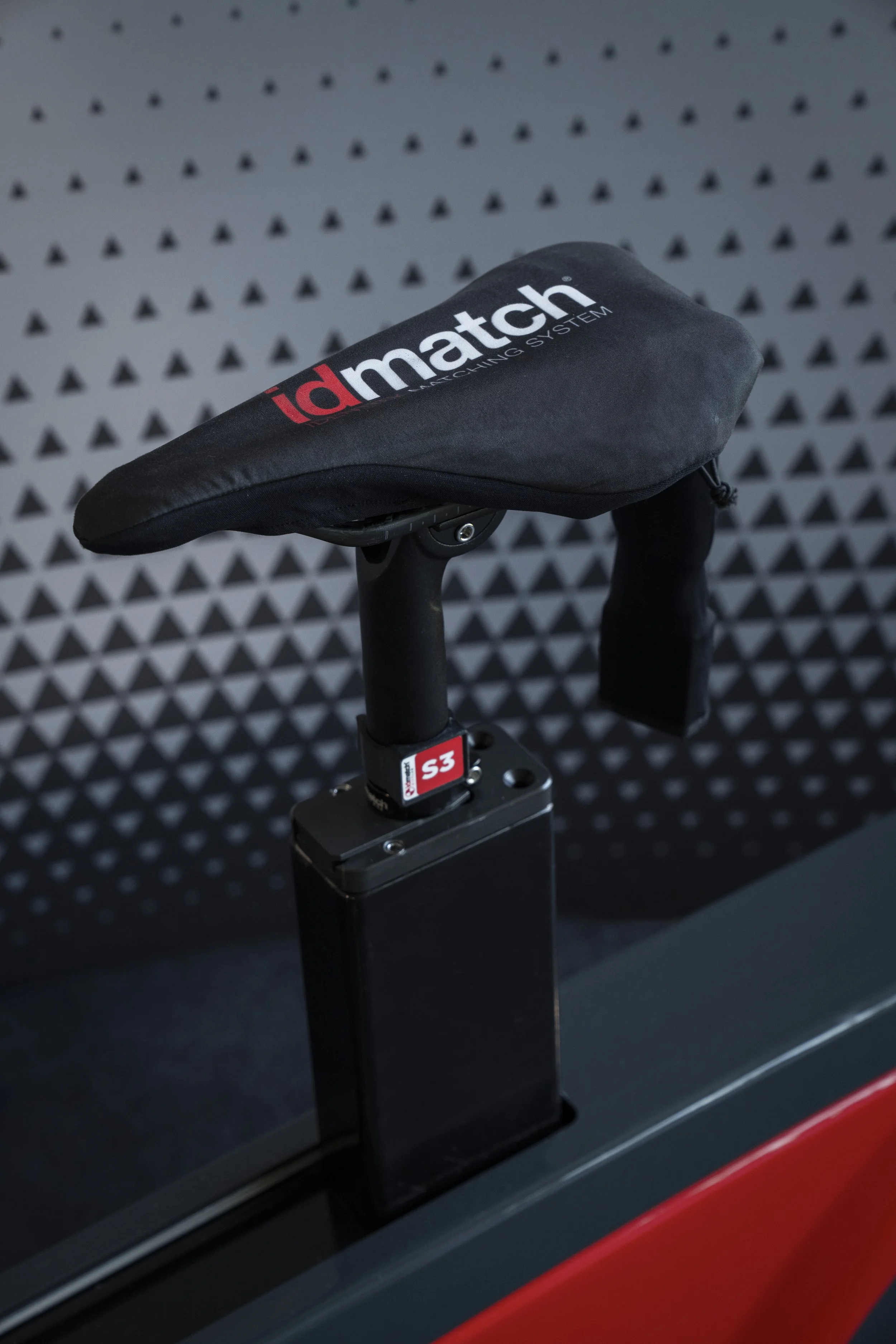 A close-up of a bicycle seat labeled 'idmatch' with a black saddle and a small red and black label on the seat post, set against a dark background with a dotted and triangular pattern.