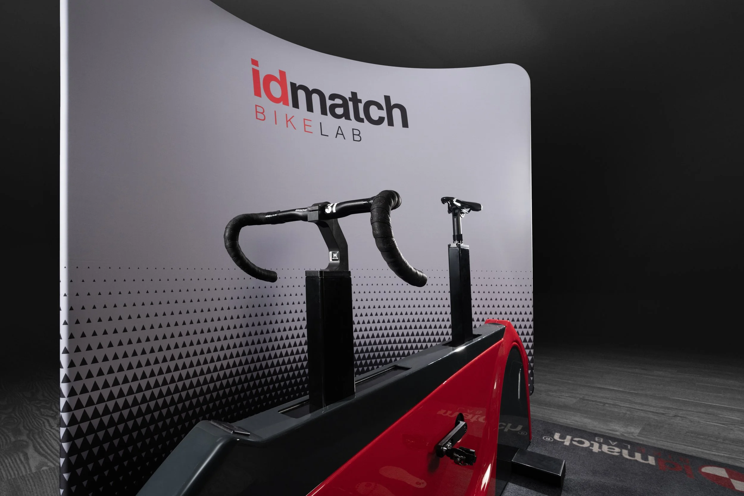 A display of a black cycling handlebar, a black saddle, and a red stationary bike or exercise bike in front of a backdrop with 'id match BIKE LAB' logo, black and white color scheme, and a patterned bottom section.