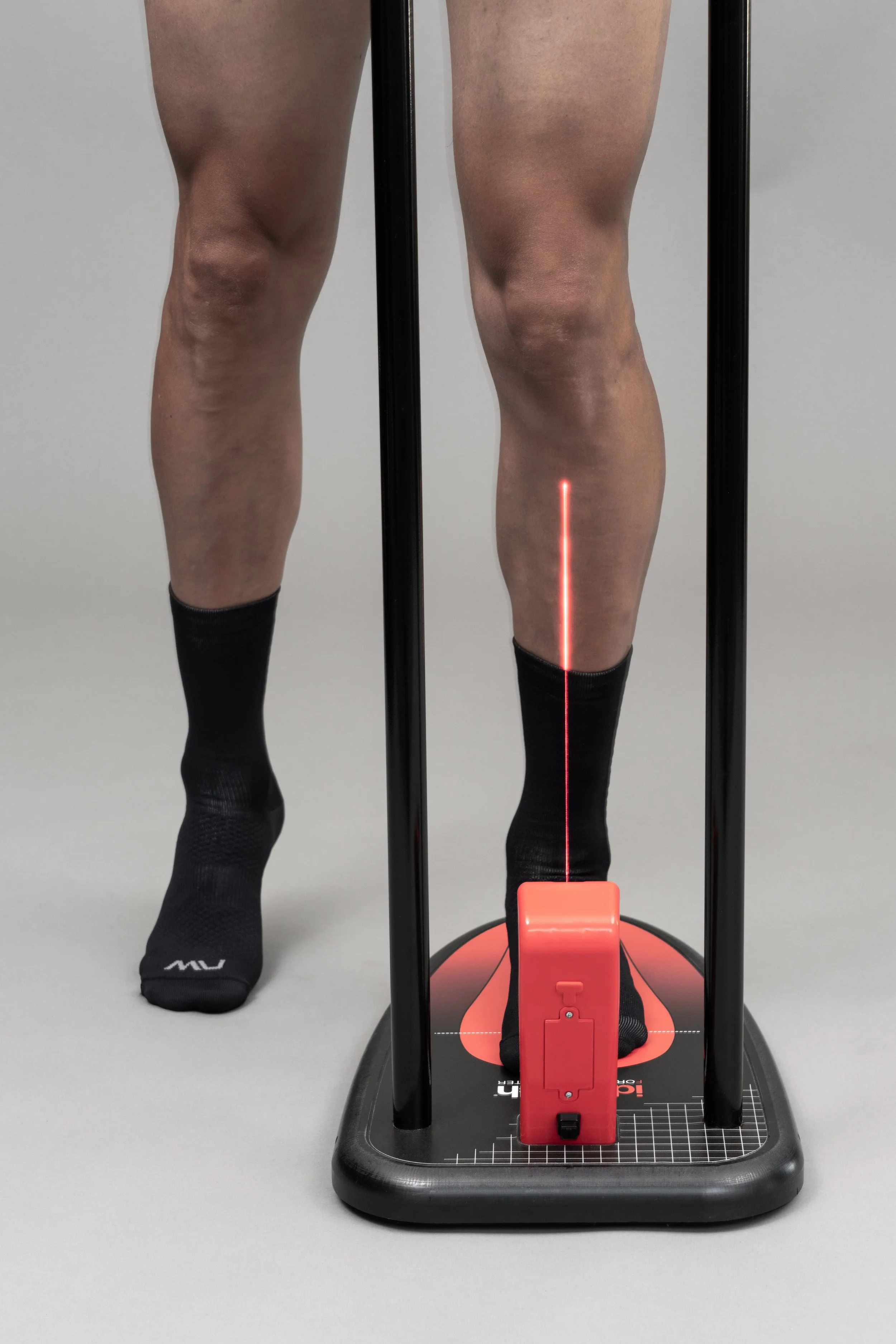 A person standing on a medical or diagnostic device with two vertical black bars, wearing black socks with the logo 'MU'. The focus is on the person's legs and feet, with the device's laser line visible on their legs.