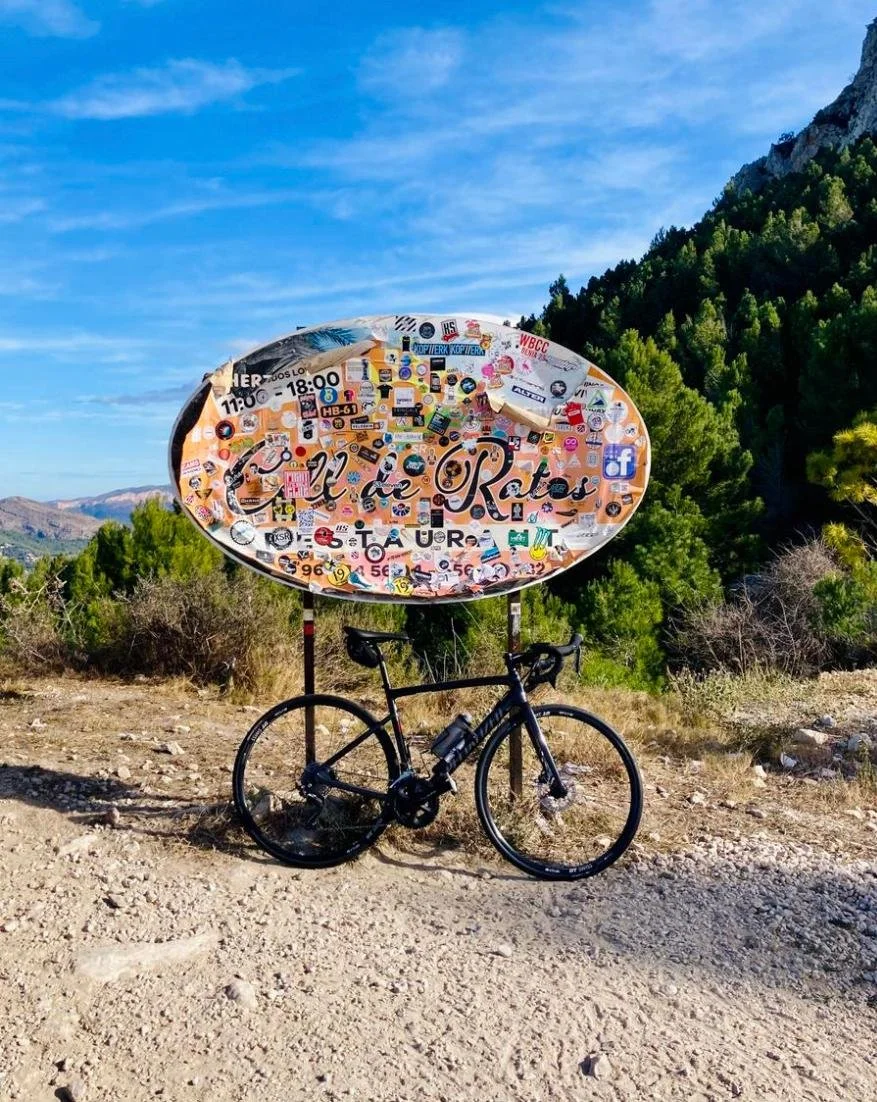 Cartel de bardas con stickers adornando la superficie, con letras que dicen 'El de Ratos', en un paisaje de montaña con un bicicleta al frente y árboles verdes al fondo, bajo un cielo azul con algunas nubes.