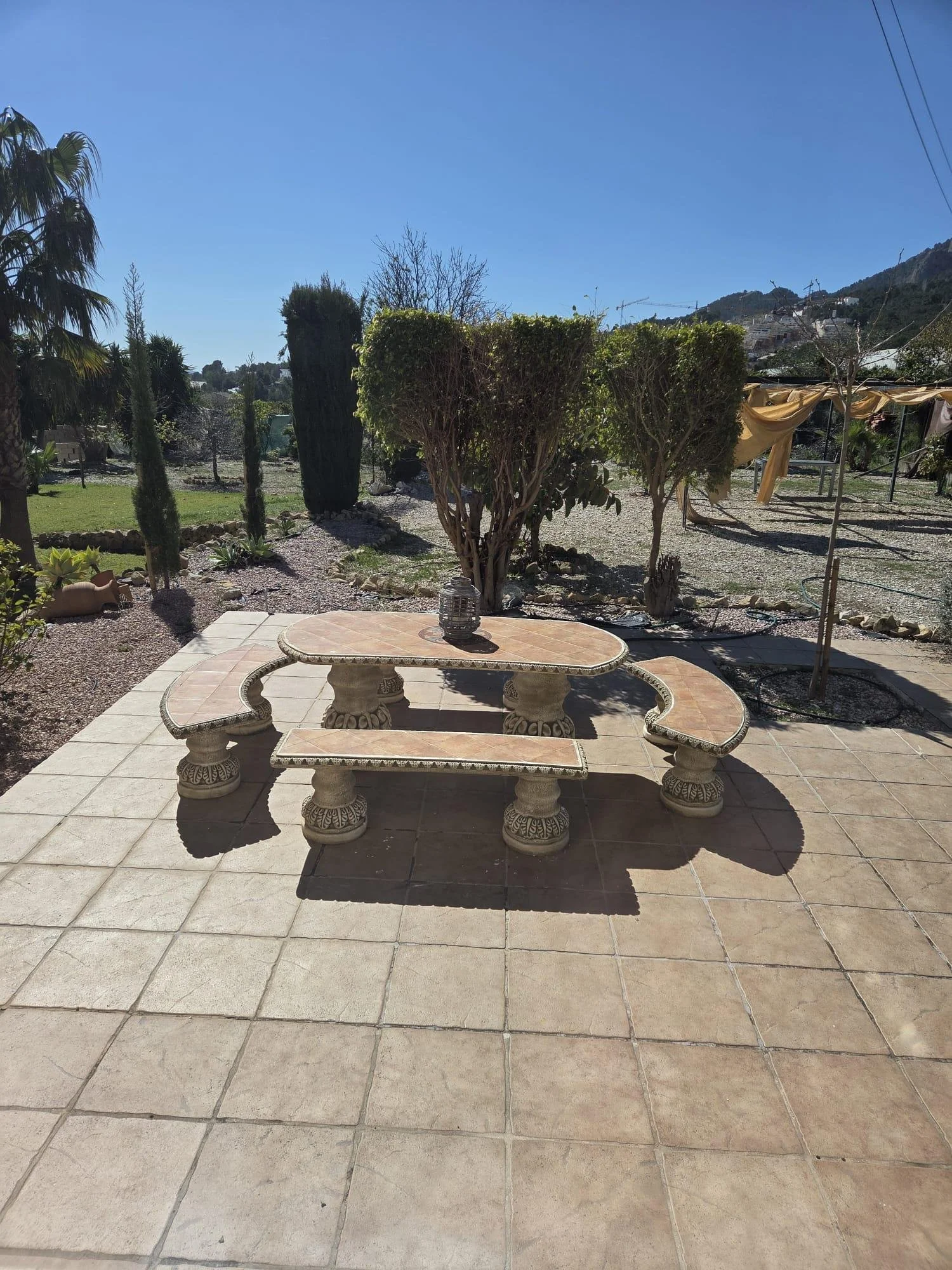 Vista de un patio exterior con mesa y bancos de piedra en primer plano, árboles y arbustos en el fondo, y colinas con casas en la distancia bajo un cielo despejado.