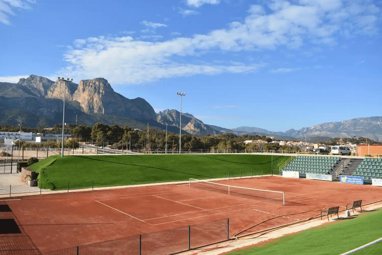 Cancha de tenis con fondo montañoso, césped y gradas en un campo de deportes al aire libre en un día soleado.