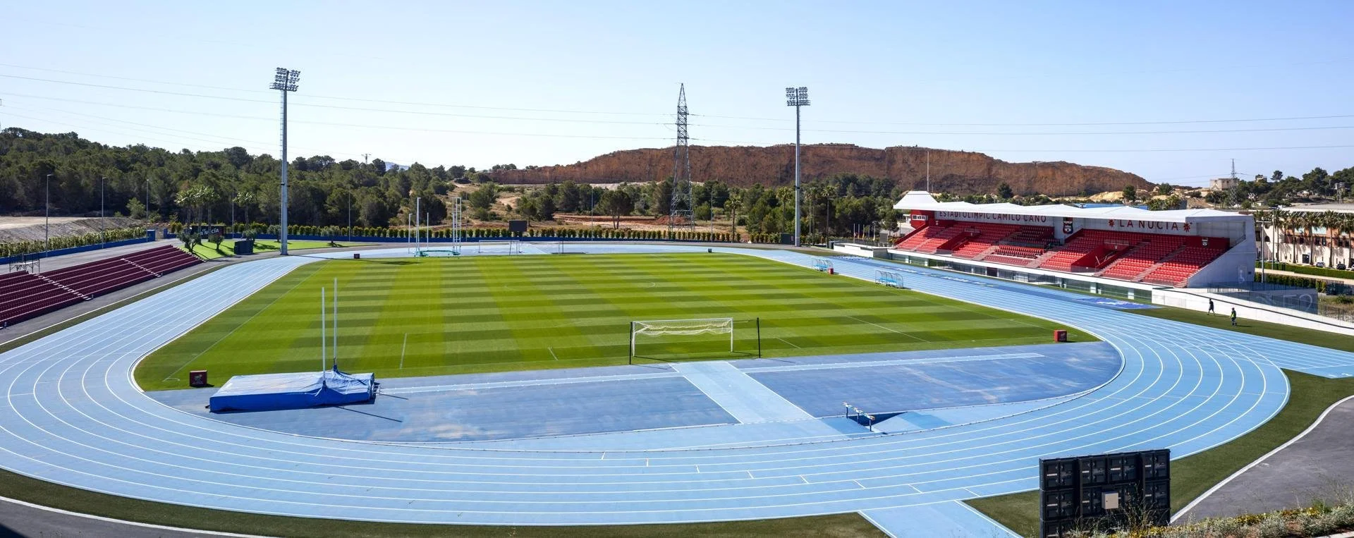 Estadio de atletismo con pista azul, campo de fútbol, graderíos y estructura de sombra, rodeado de naturaleza y colinas en el fondo.