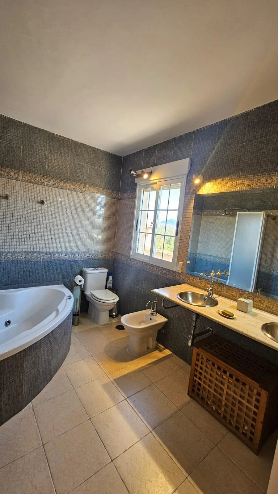 Baño con ducha, inodoro, bidé, lavabo con consola de mármol, espejo grande, ventana con rejas y vista a campo, azulejos de tonos grises y beige, piso de baldosas beige.