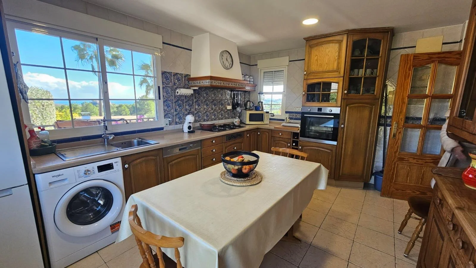 Cocina de madera con gabinetes de madera, horno empotrado, microondas, estufa con campana, lavavajillas, lavadora, mesa rectangular con mantel y fruta en un recipiente en el centro, ventanas grandes con vista a un jardín y árboles, piso de baldosas beige.