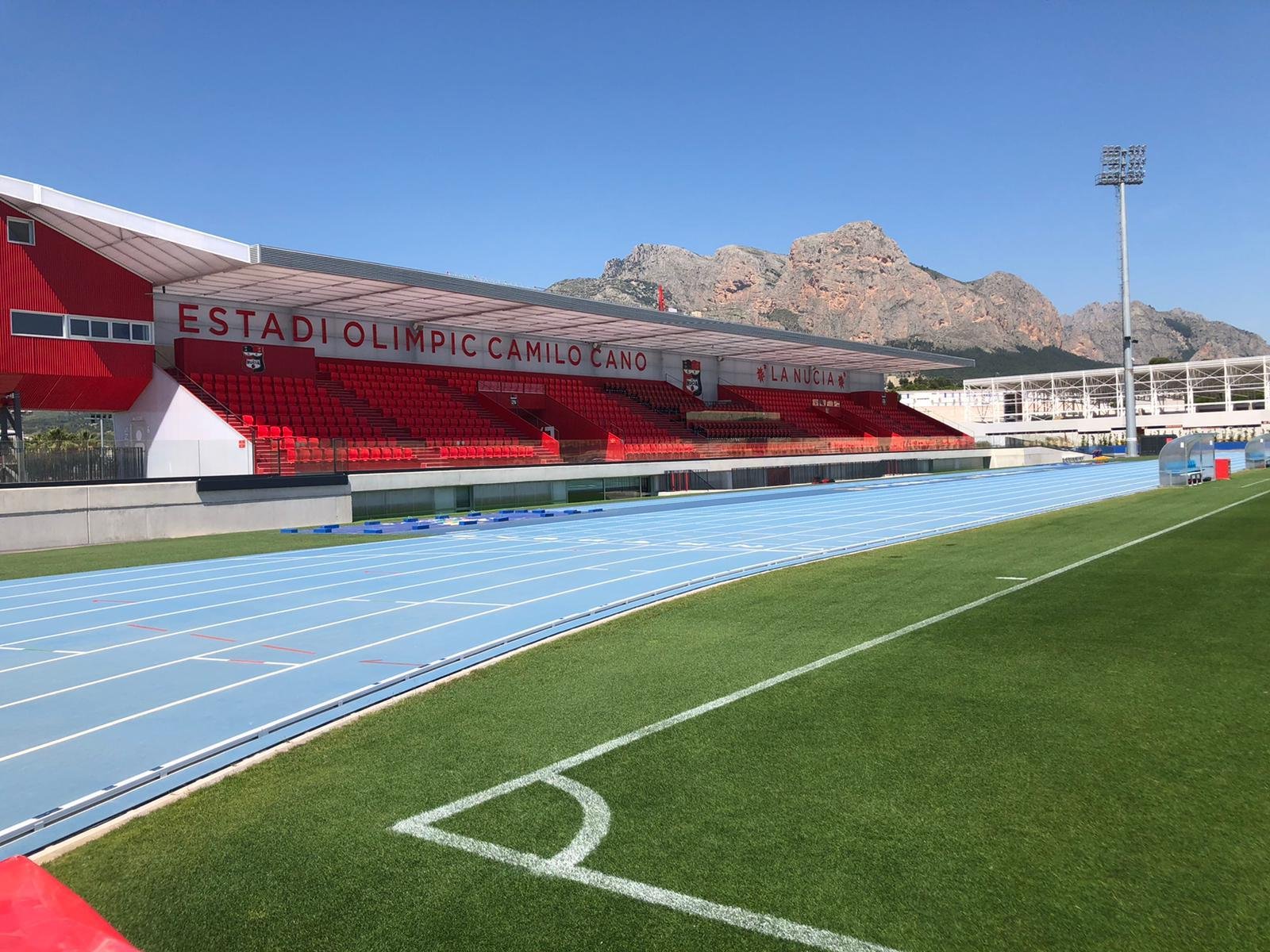 Estadio olímpico con gradería roja, campo de atletismo azul y montañas al fondo bajo un cielo claro.