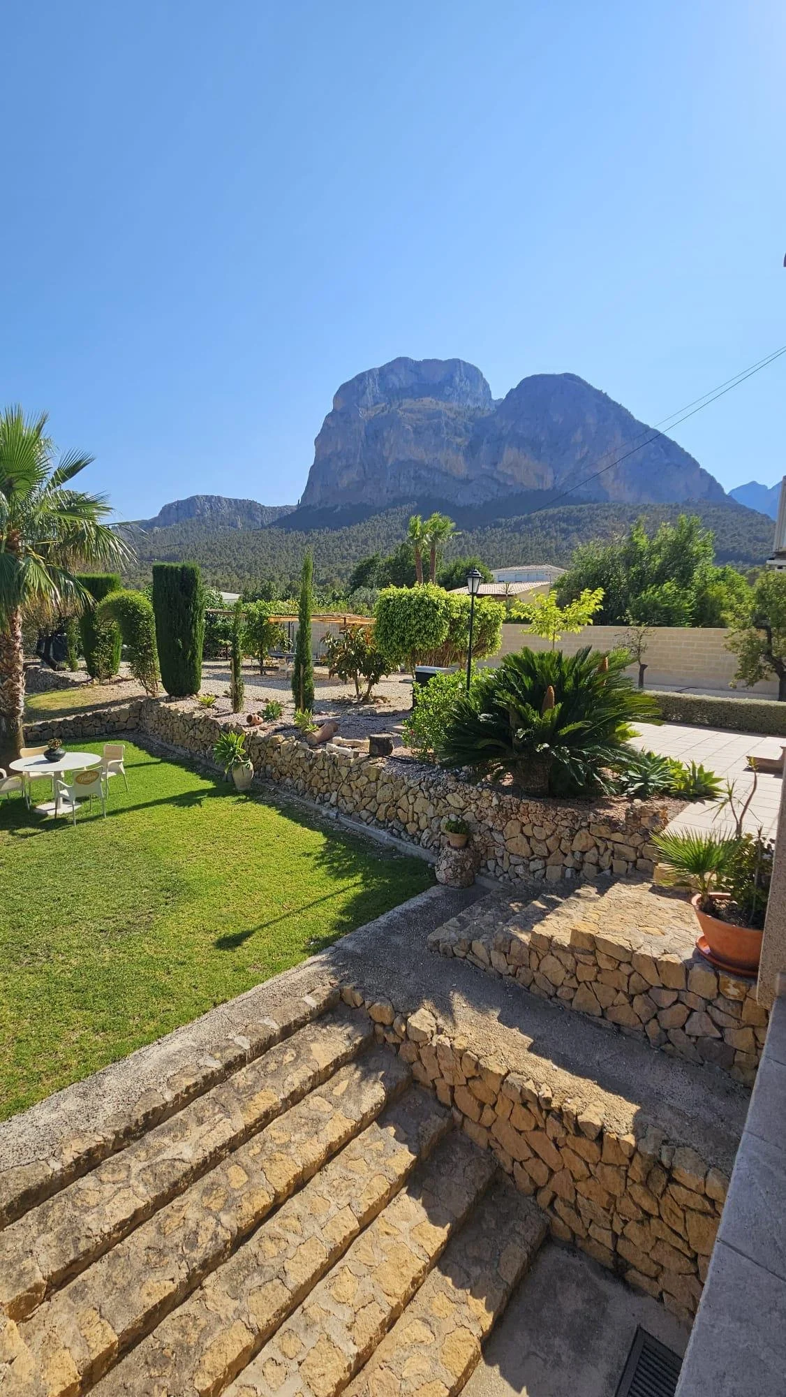 Jardín con césped, plantas, macetas, escalones de piedra, y montañas en el fondo bajo un cielo claro.