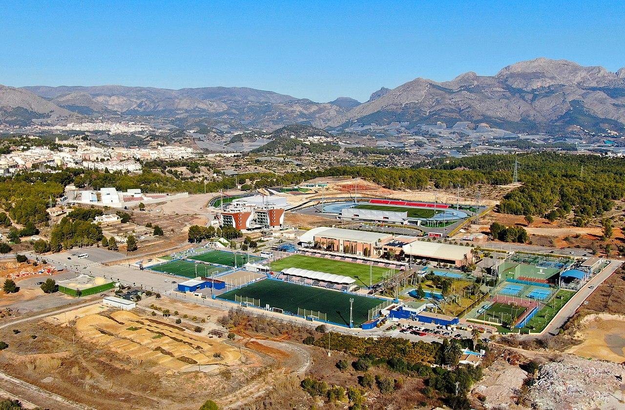 Vista aérea de instalaciones deportivas con canchas de fútbol, tenis y otros deportes, rodeadas de áreas verdes y montañas en el fondo.