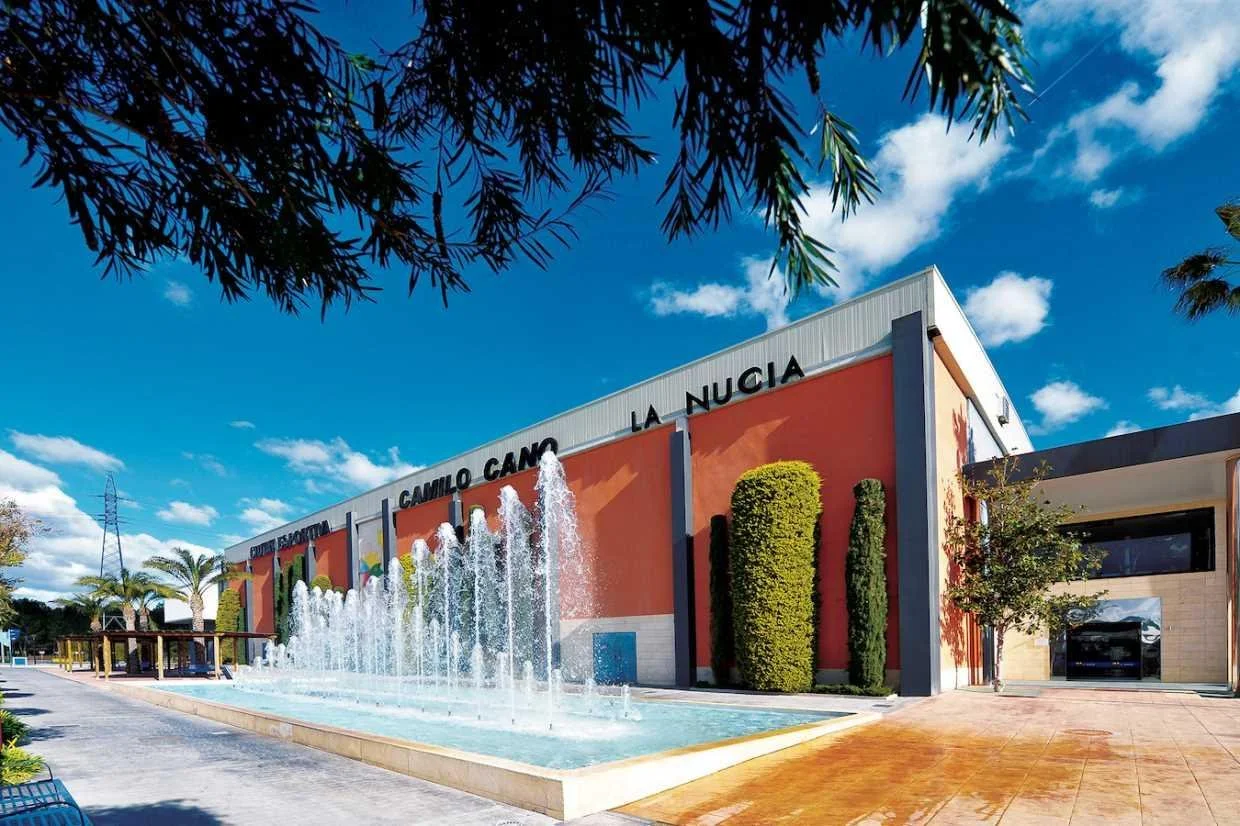Fachada de un edificio de color naranja con letras negras que dice 'CAMILO CANO LA NUCIA', con fuente de agua en la entrada, árboles y césped alrededor, cielo azul con algunas nubes.