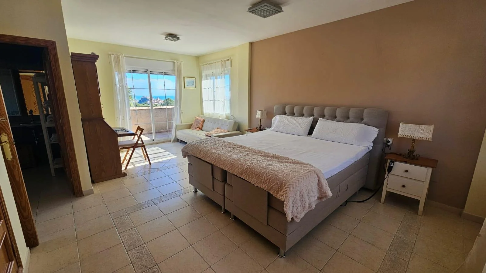 Habitación con cama grande, sofá, mesas de noche, vista al mar a través de ventanas y puerta corrediza hacia balcón, decoración clara y sencilla.