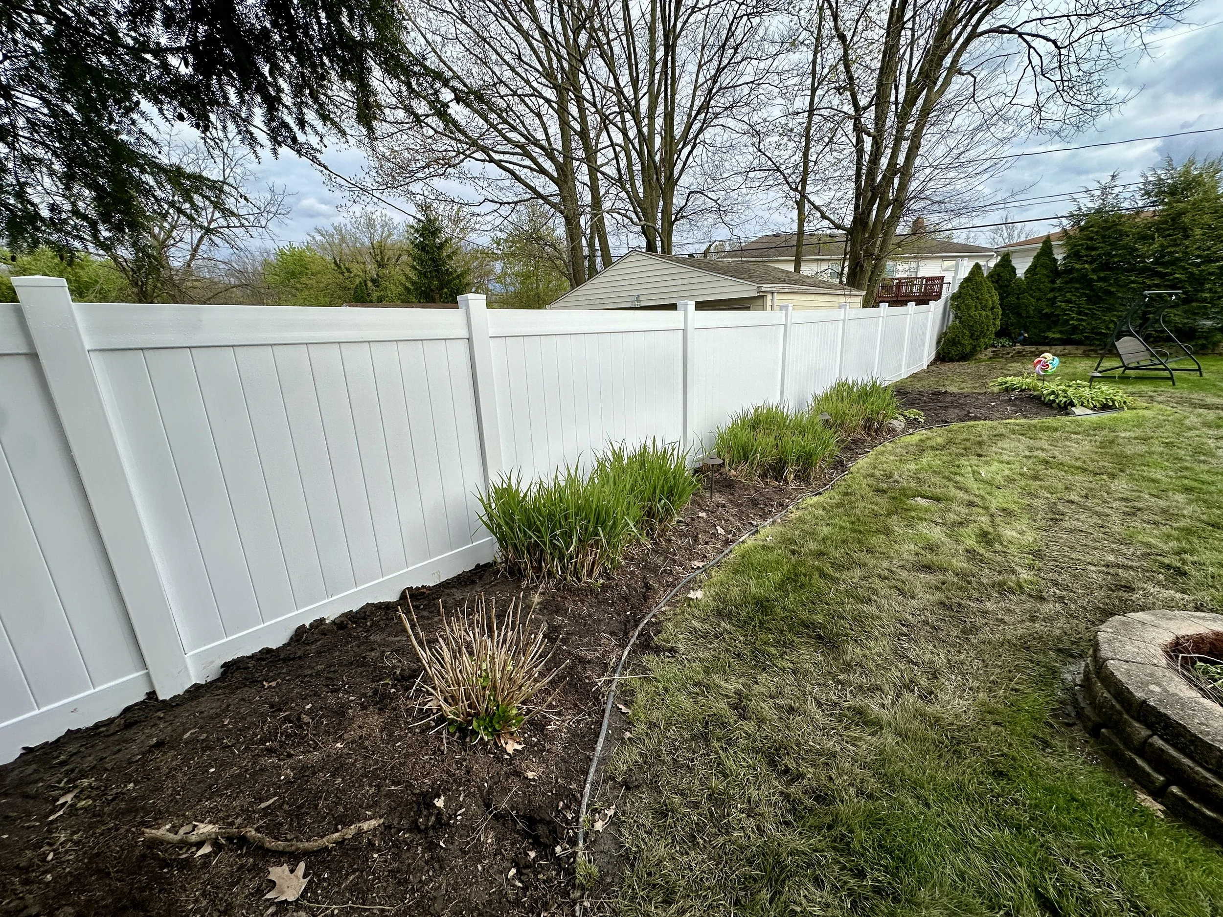 A backyard with a white vinyl fence, a garden bed with young plants, and a lawn with a garden chair and a colorful pinwheel. There are large trees with bare branches and a neighboring house with a deck in the background. The sky is cloudy.