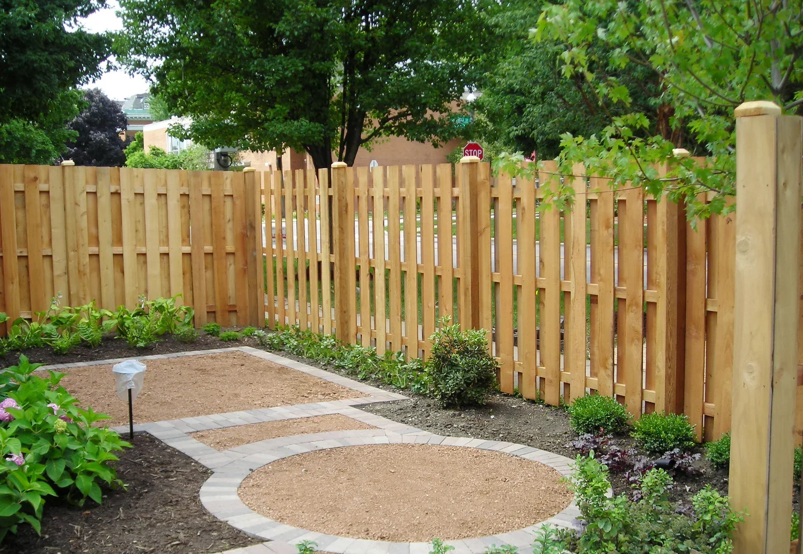 A backyard garden with a wooden fence, small plants, flower bed, and a gravel pathway with a circular design.