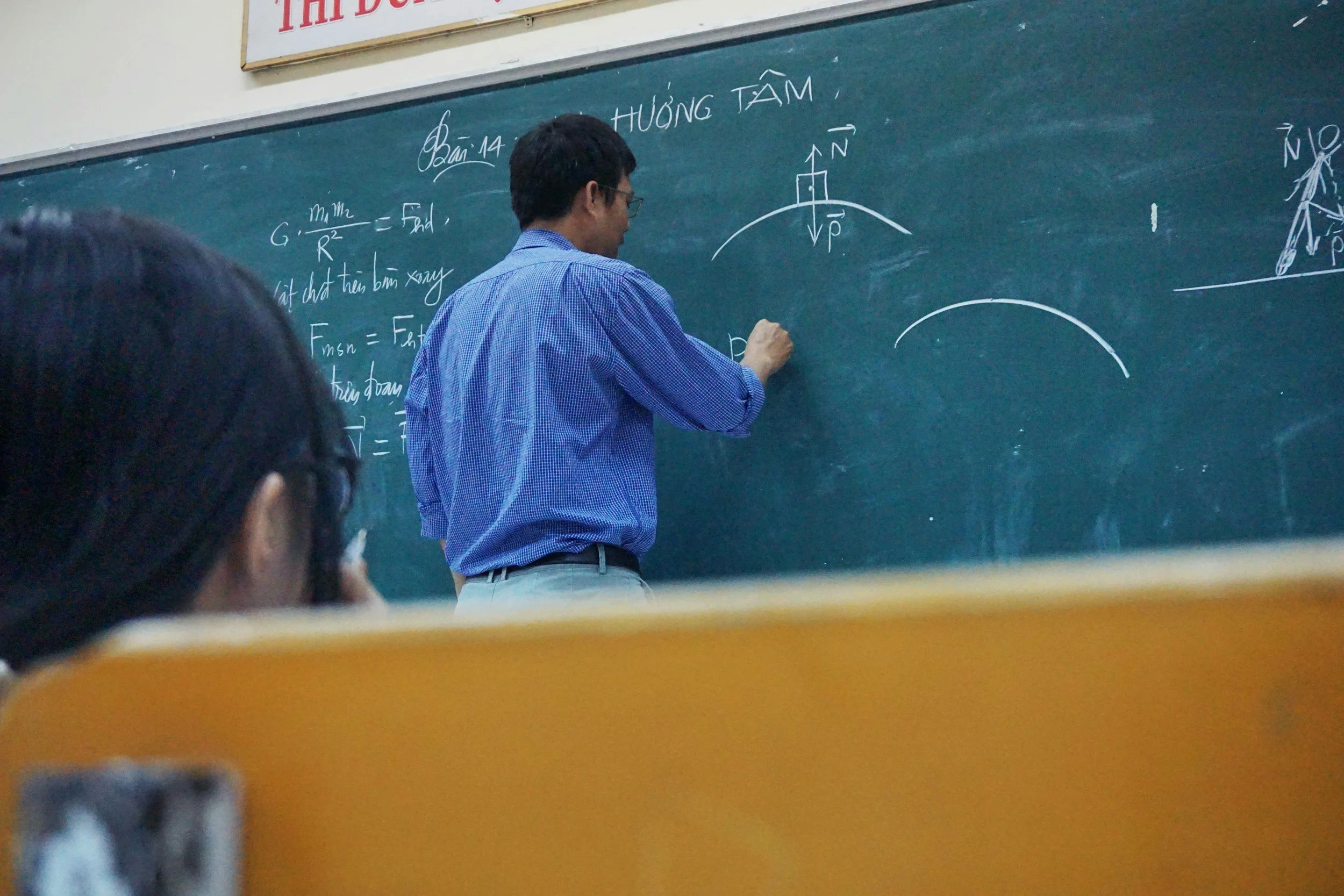 A teacher standing at the blackboard, writing with chalk in a classroom, with students in the foreground.