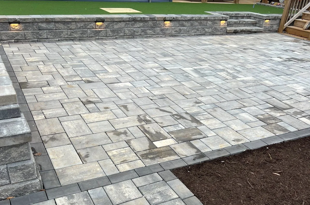 A newly paved outdoor area with gray stone bricks arranged in a pattern, bordered by darker bricks, and a small section of dark soil at the bottom right corner. In the background, there's a retaining wall with built-in lighting, and a small set of st