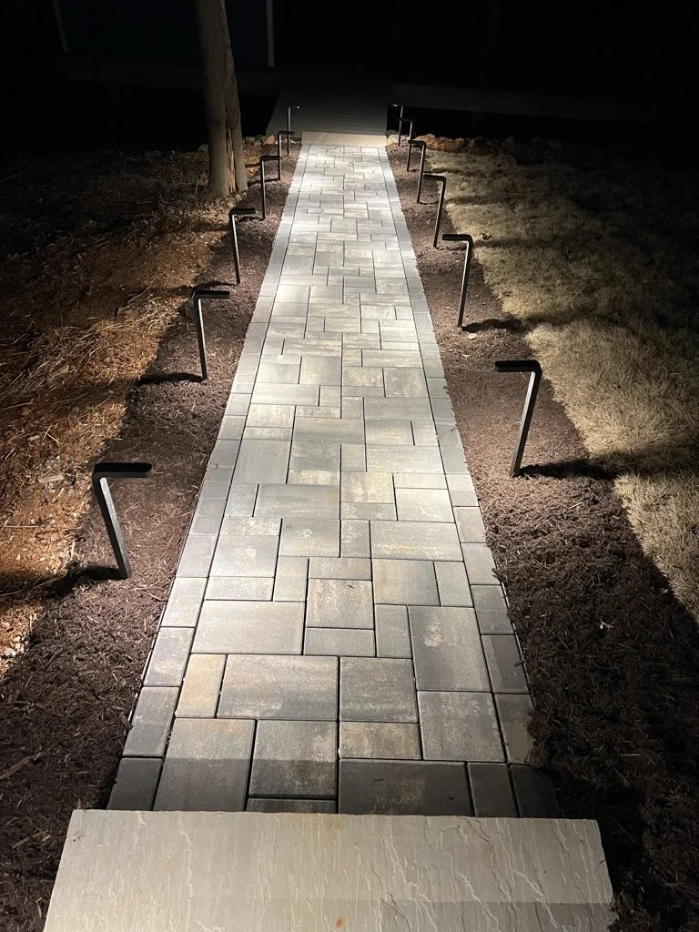 Nighttime view of a paved walkway made of gray rectangular bricks, with black metal guardrails on both sides, surrounded by dirt and grass.