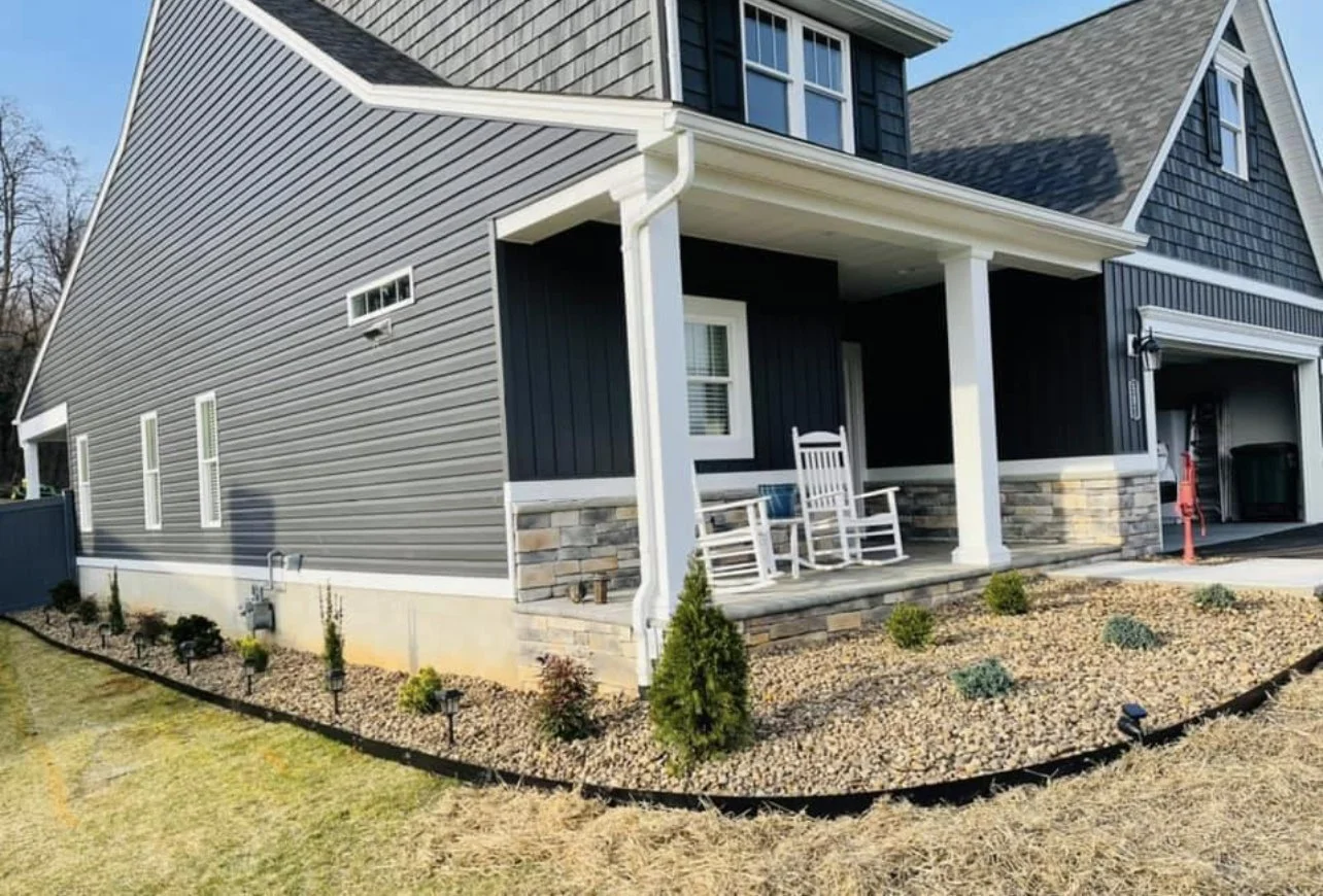 A modern house with dark gray vinyl siding, stone accents, a front porch with white rocking chairs, and a landscaped yard with small plants and mulch.