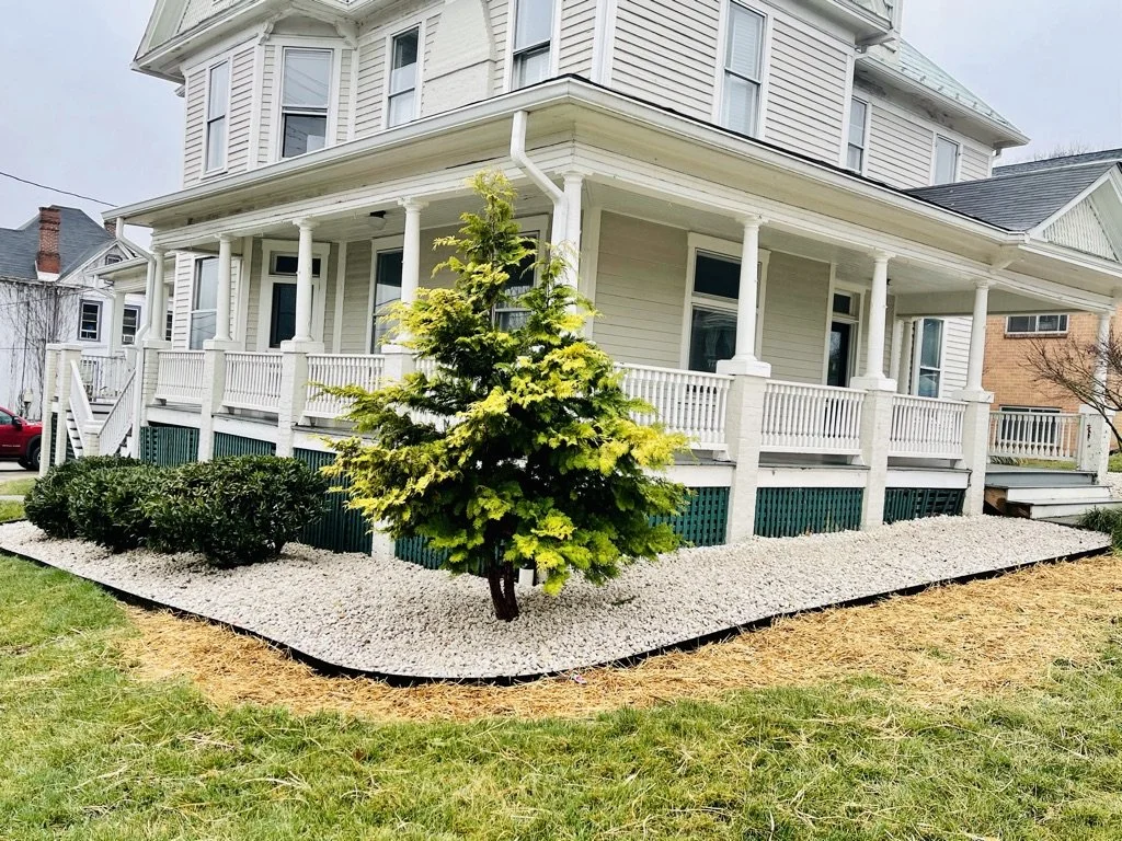 A large white two-story house with a wrap-around porch, white railings, and multiple windows. The yard has a small landscaped area with a pine tree, some bushes, white gravel, and a grassy lawn.