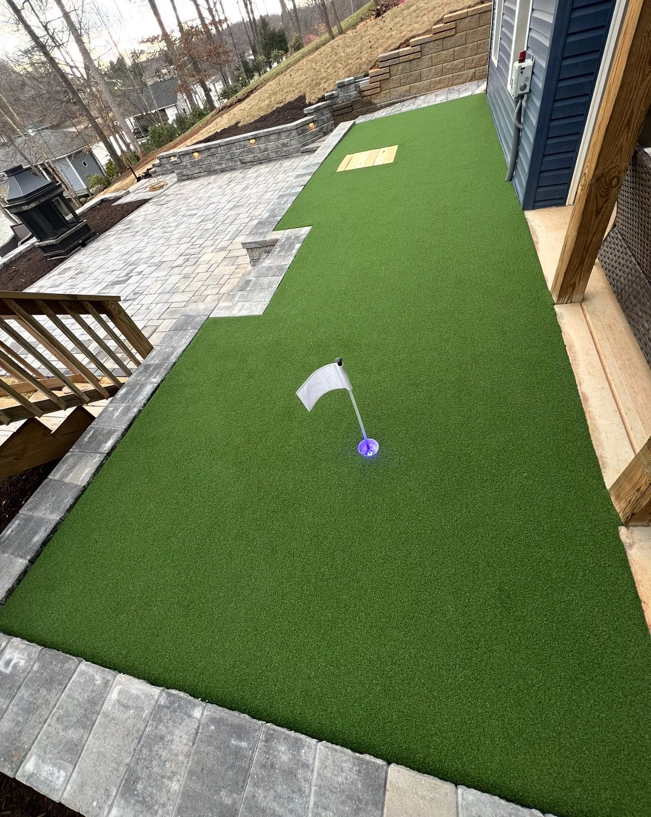 Artificial turf putting green with a white flag and lit base for night play, near a patio with stone pavers and a brick wall for landscaping.