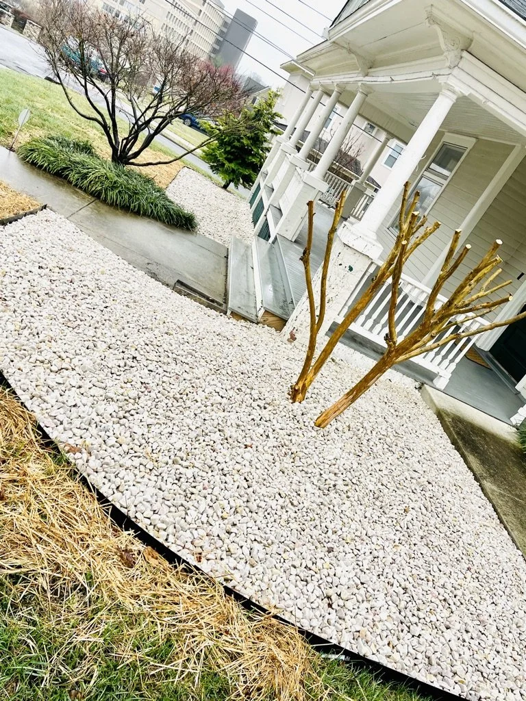Front yard with gravel landscaping, pruning new trees, and a porch on a cloudy day.