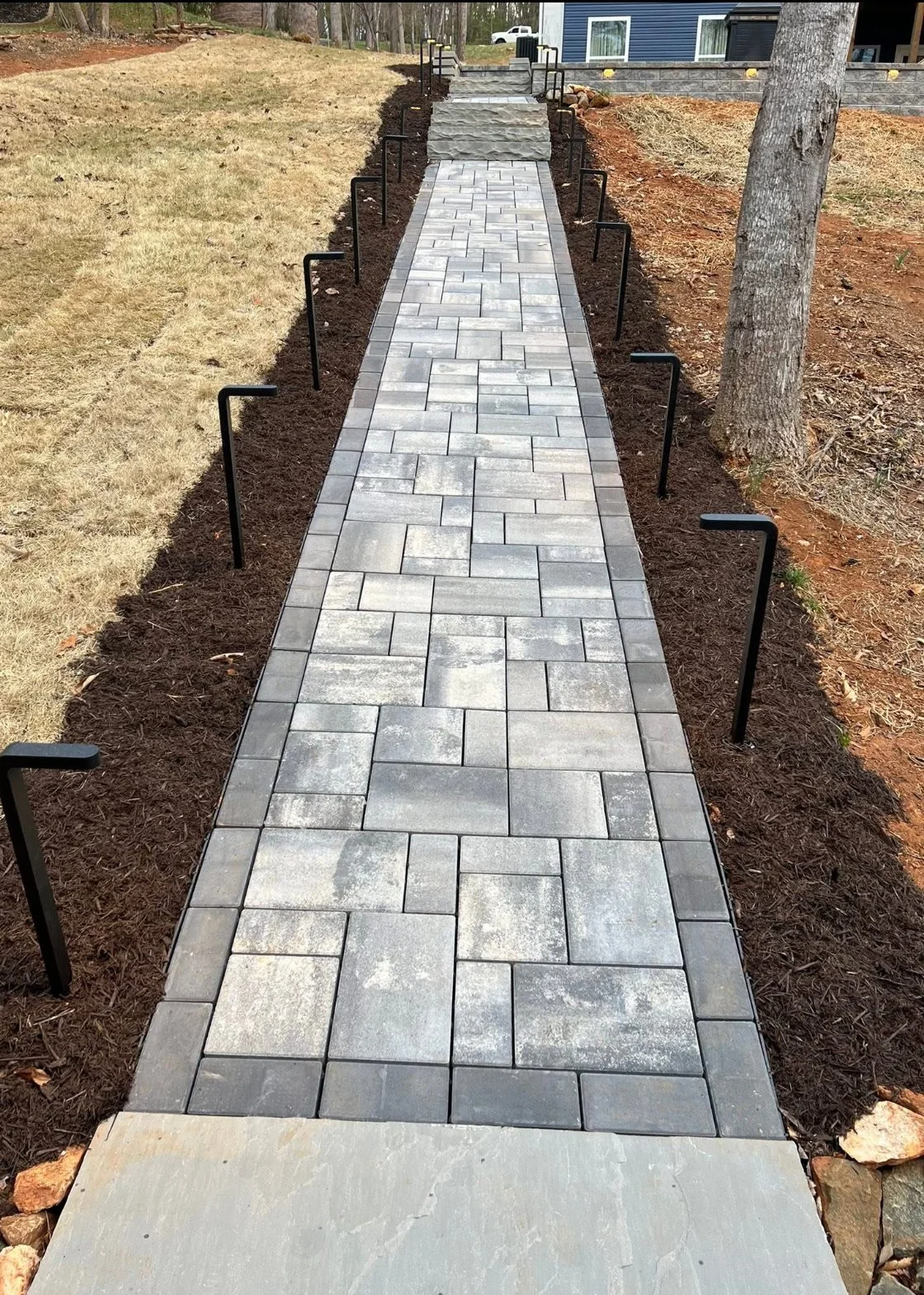 Constructed walkway with gray paving stones, bordered by dark soil and soil-covered patches, with black metal stakes on either side, leading up to stone steps near a house with blue siding, surrounded by trees and yard.