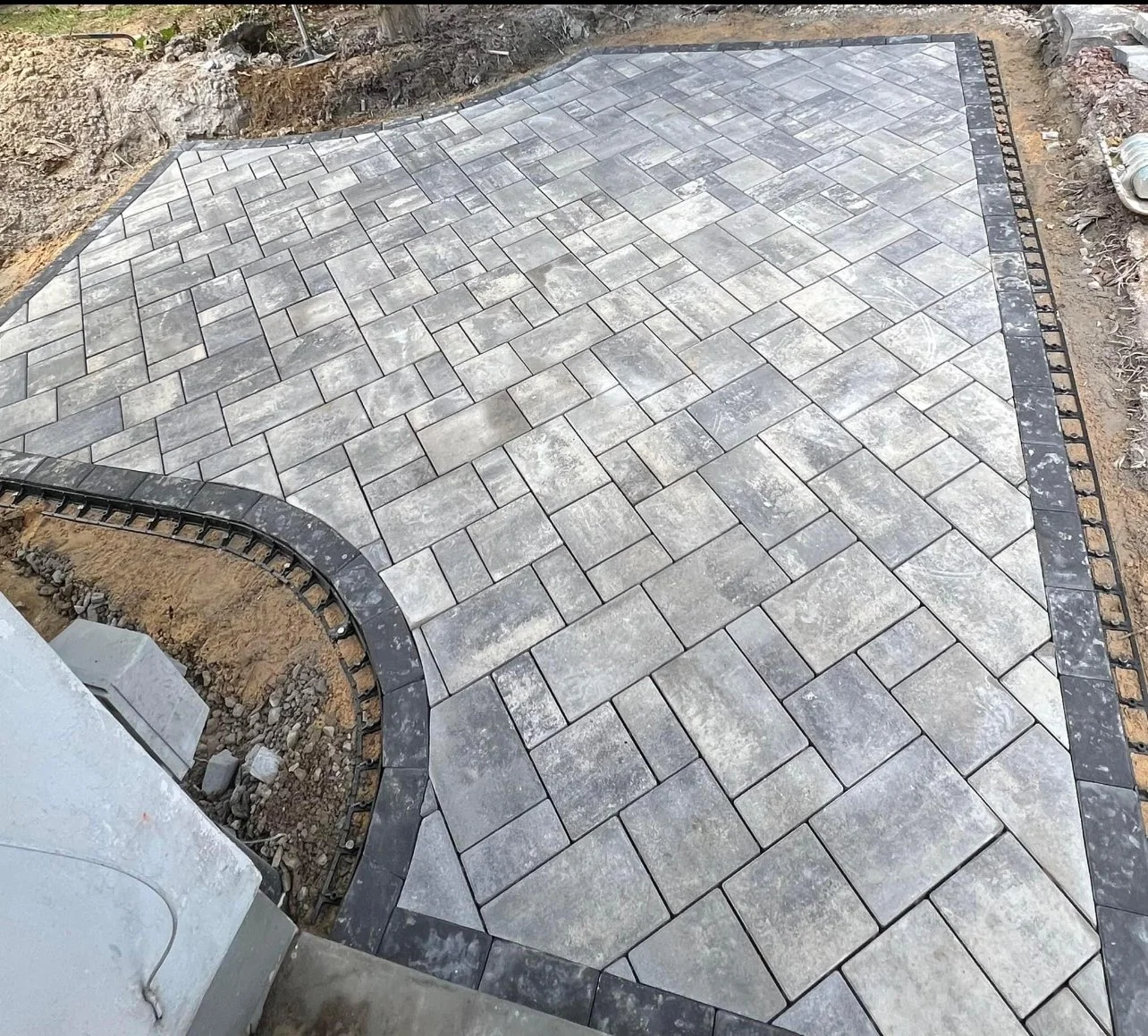 Newly paved stone patio with gray and black stones, curved edges, and a border of darker stones, under construction with exposed dirt and construction materials nearby.