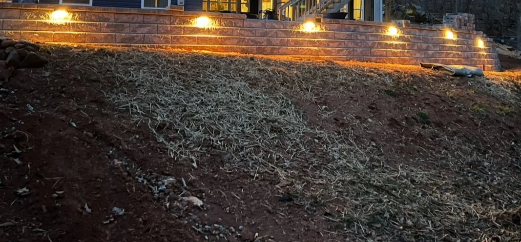 Slope of bare soil with dry grass and rocks at the bottom, illuminated by outdoor lights at the top of the slope.