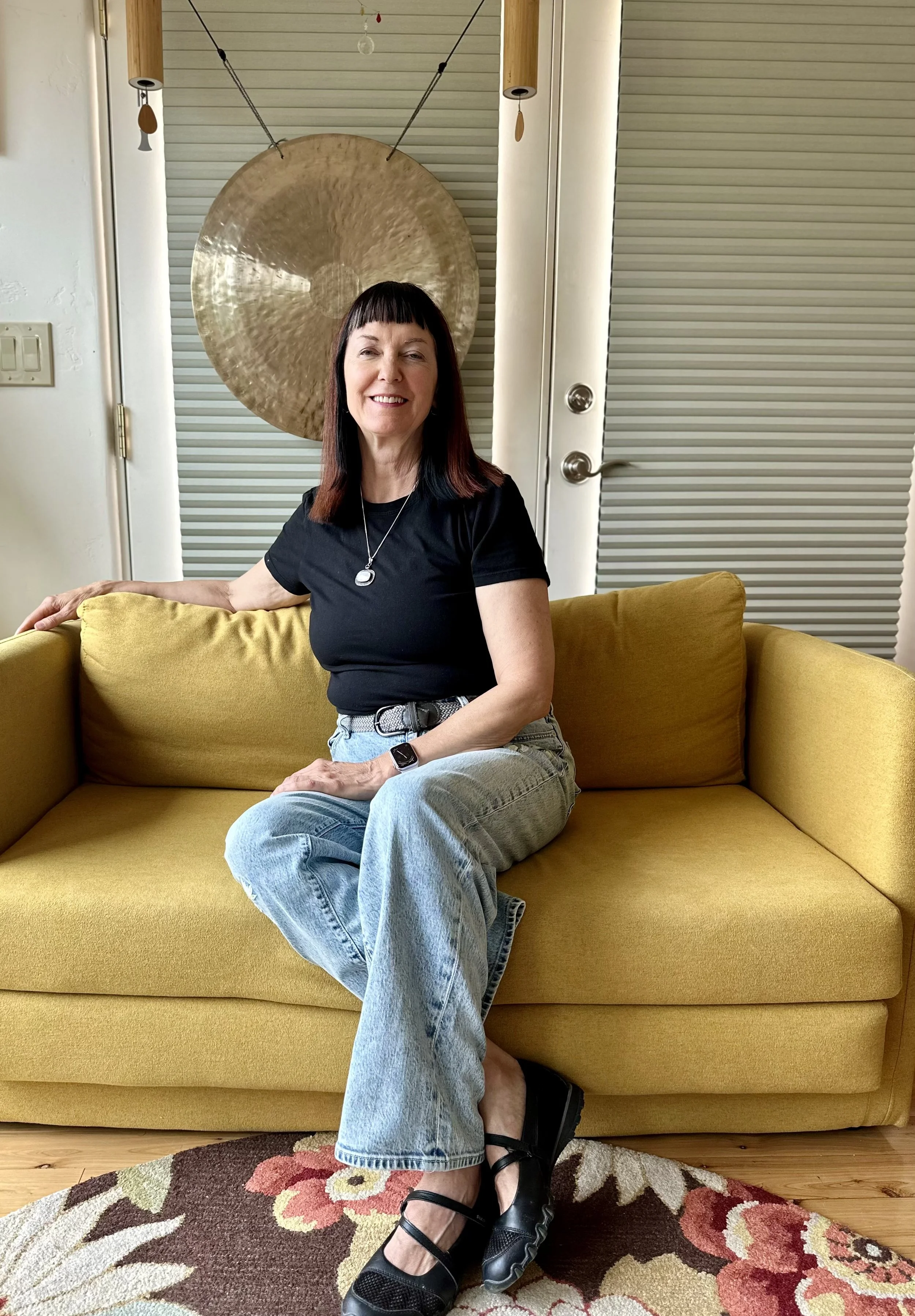 A woman with dark brown hair and bangs smiling, sitting on a yellow couch in a room with beige walls and a large golden gong behind her. She is wearing a black t-shirt, light blue jeans, black sandals, and a watch on her left wrist.