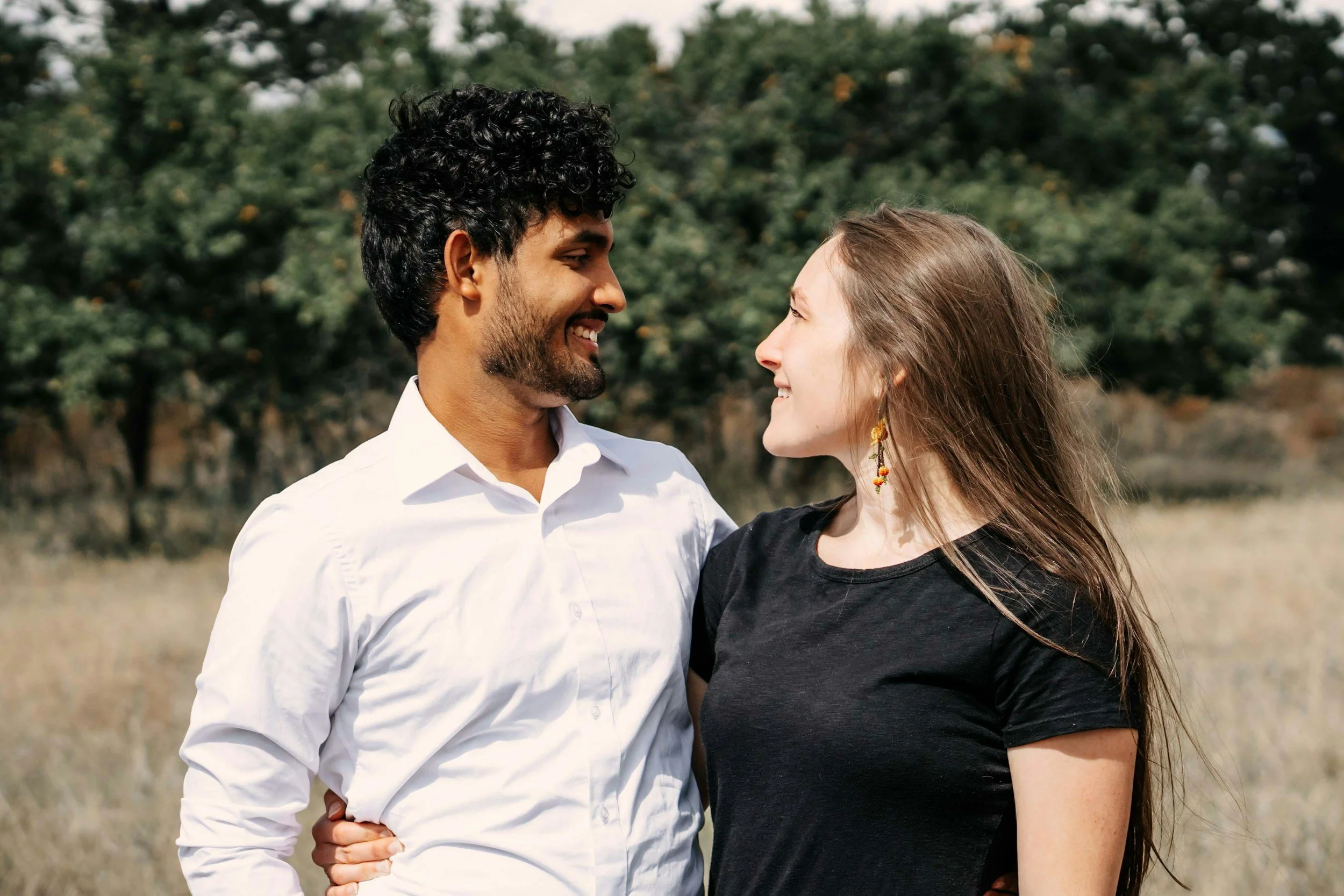 A man and woman smiling at each other outdoors with trees in the background.