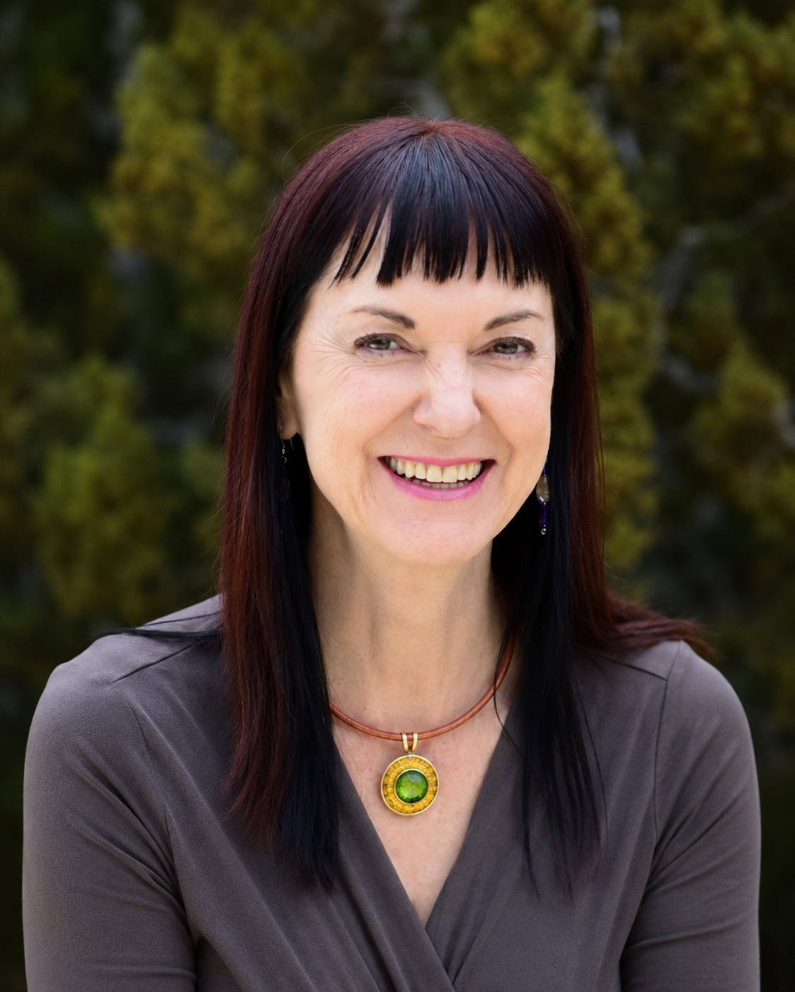 A woman with dark red and black hair smiling outdoors, wearing a gray top and a colorful pendant necklace, with blurred green foliage in the background.