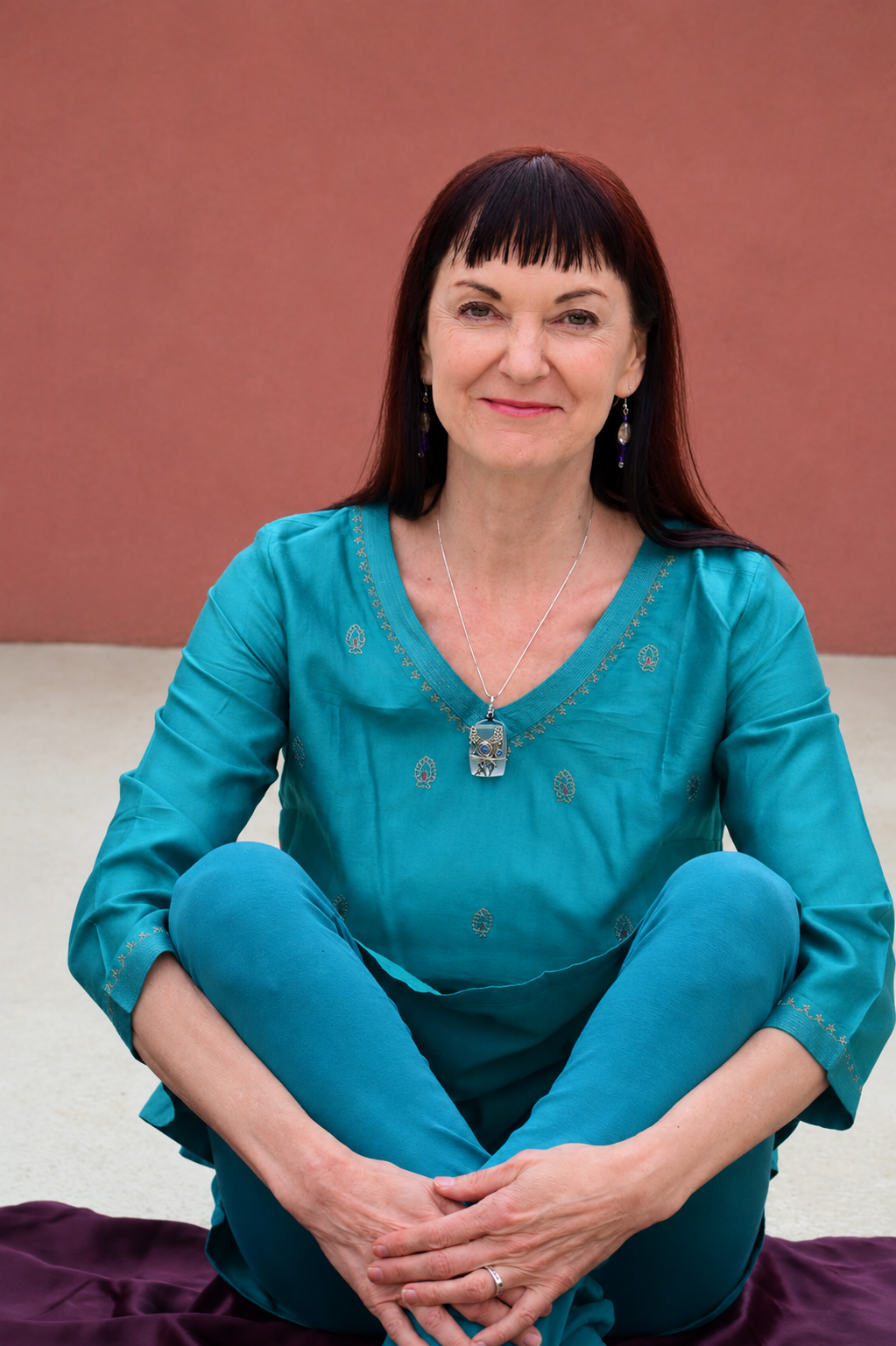A woman with dark hair and bangs sitting cross-legged on a purple cloth, wearing a teal outfit and jewelry, smiling at the camera against a reddish-brown wall background.
