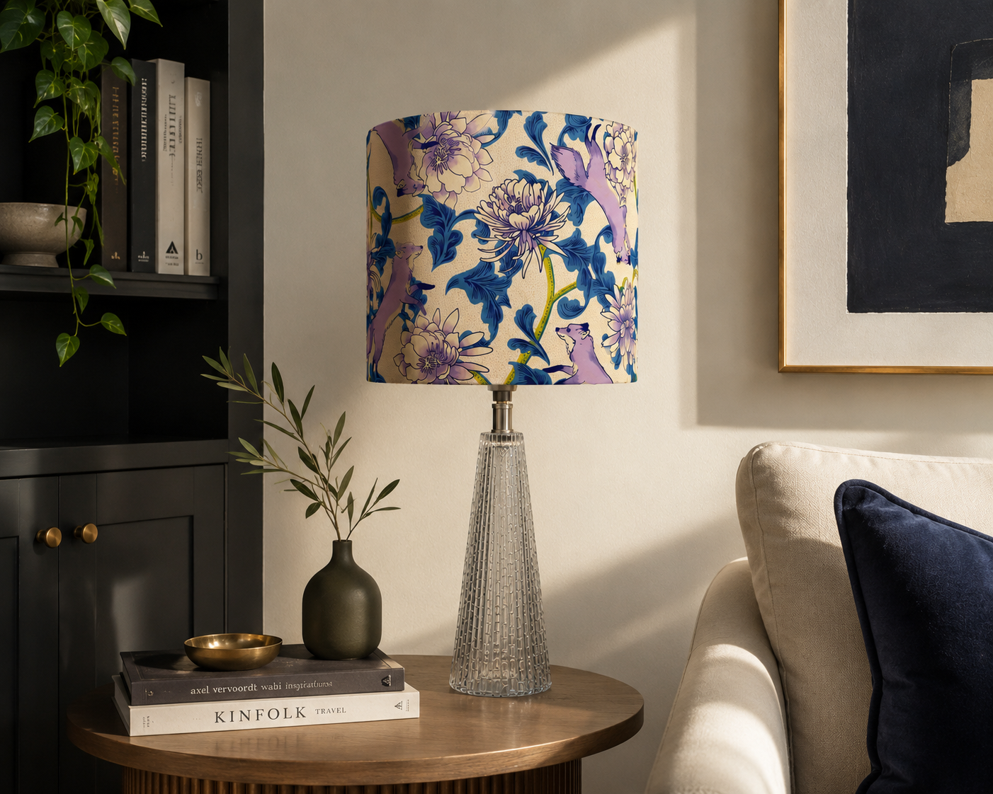 A table lamp with a floral-patterned shade featuring purple and blue flowers and animals, on a wooden side table next to a beige sofa with a navy blue pillow, a green vase with branches, and a few books.
