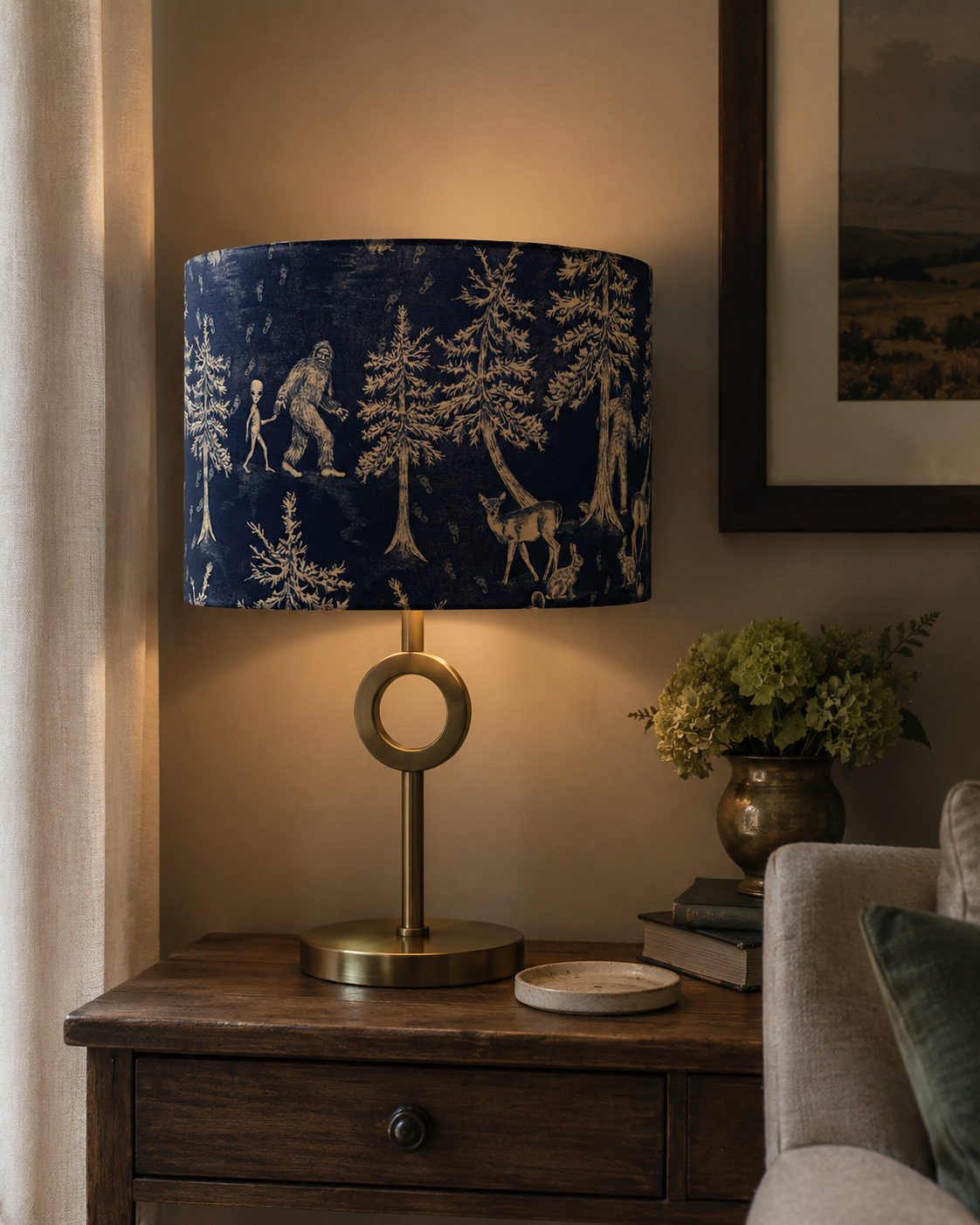 A table lamp with a blue shade featuring forest and animal motifs, placed on a wooden side table with a brass circle base, next to a vase of green hydrangeas, books, and a beige sofa.