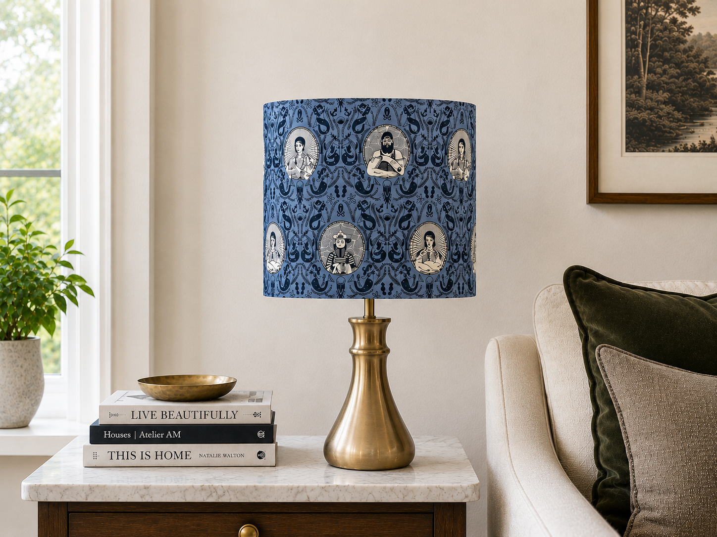 A living room side table with a gold lamp featuring a patterned blue shade with illustrated faces, a stack of books, a small decorative bowl, and a nearby armchair with pillows, with a potted plant and a window in the background.