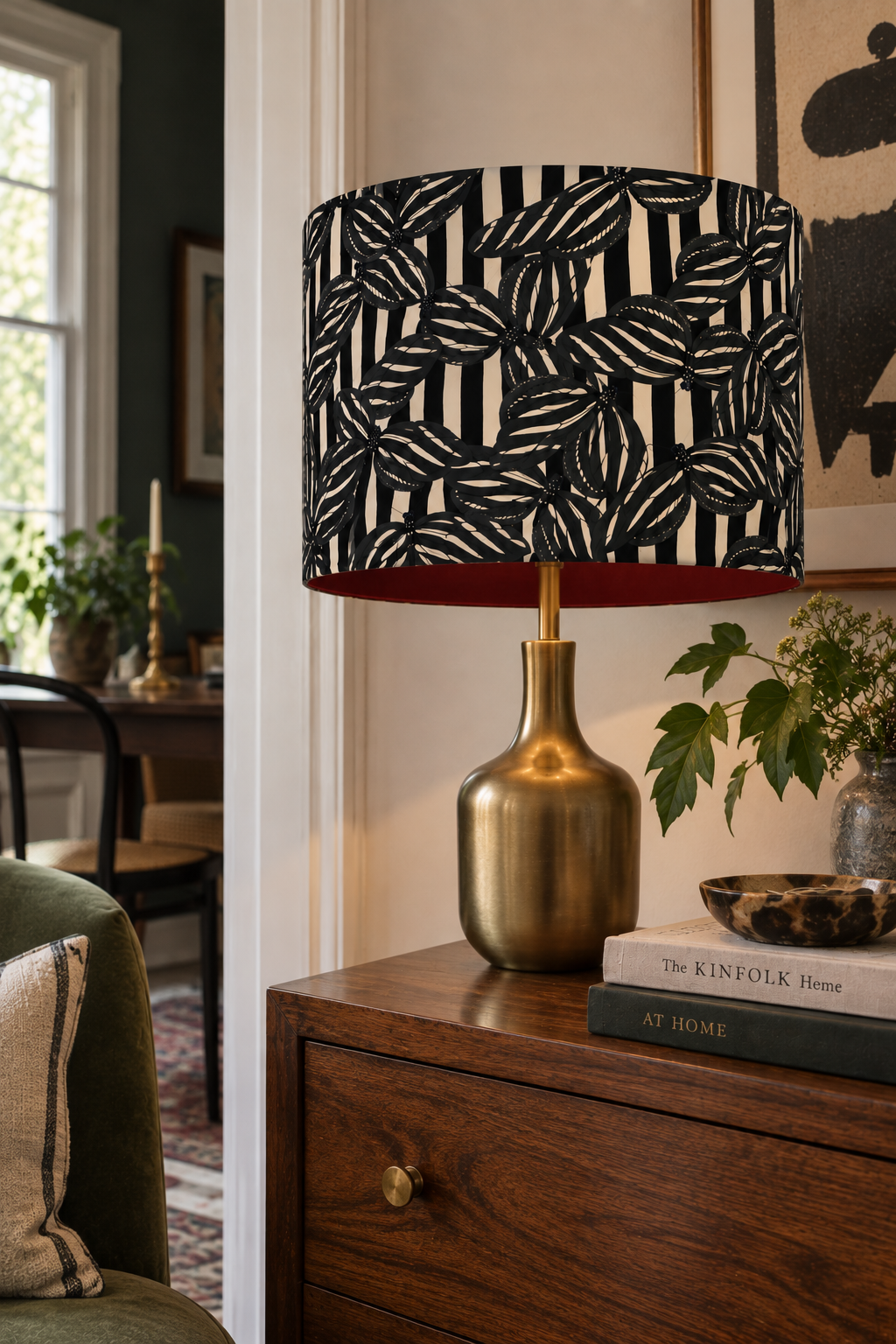 A table lamp with a black and white floral patterned lampshade on a wooden side table. The table also has a potted plant, a decorative bowl, and stacked books.