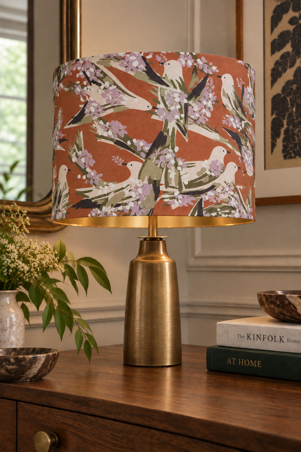 Table lamp with a floral and bird patterned shade on a wooden surface, with a small bowl, a white potted plant, and stacked books nearby.